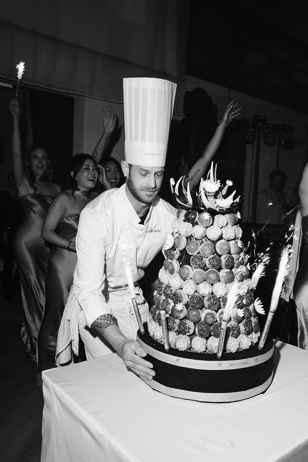 Black and white shot of chef presenting sparkler-lit croquembouche as guests dance at reception