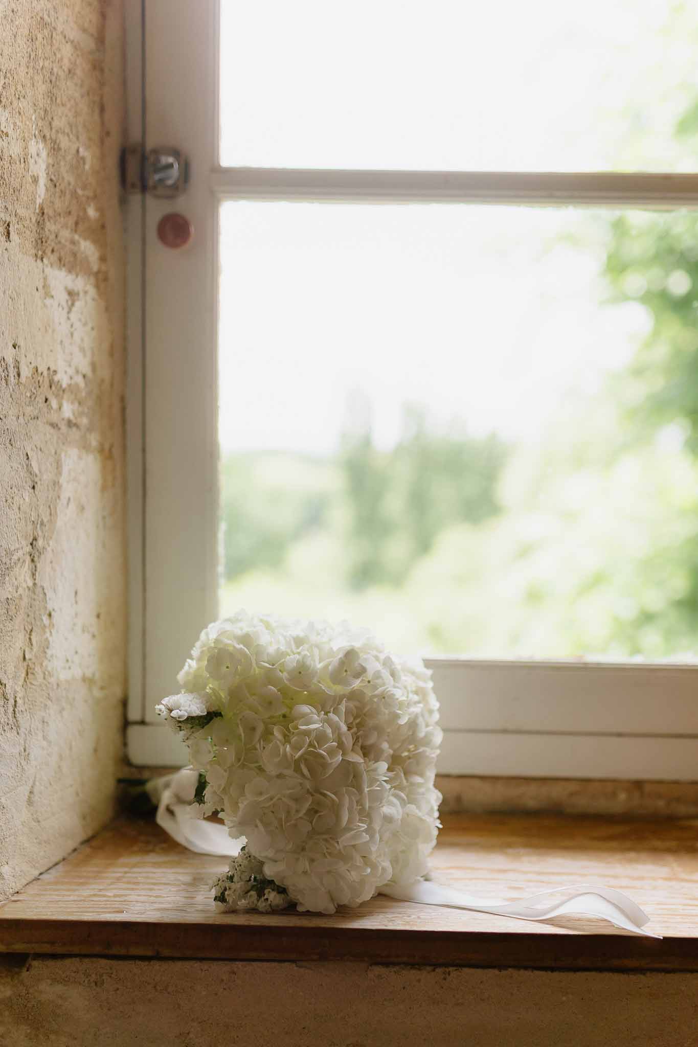 All-white hydrangea bridal bouquet with satin ribbon on wooden windowsill backlit by natural light in stone building