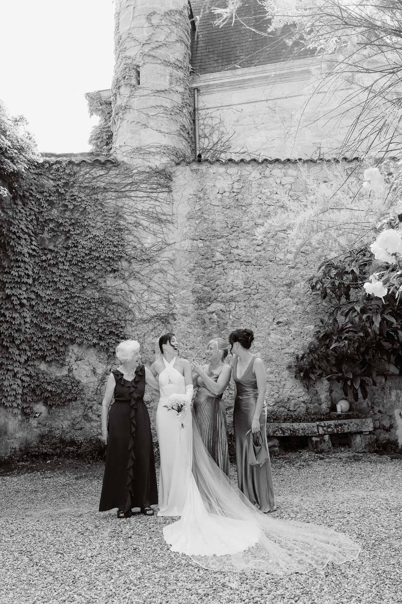Black and white candid of bride with three companions laughing at ivy-covered chateau tower