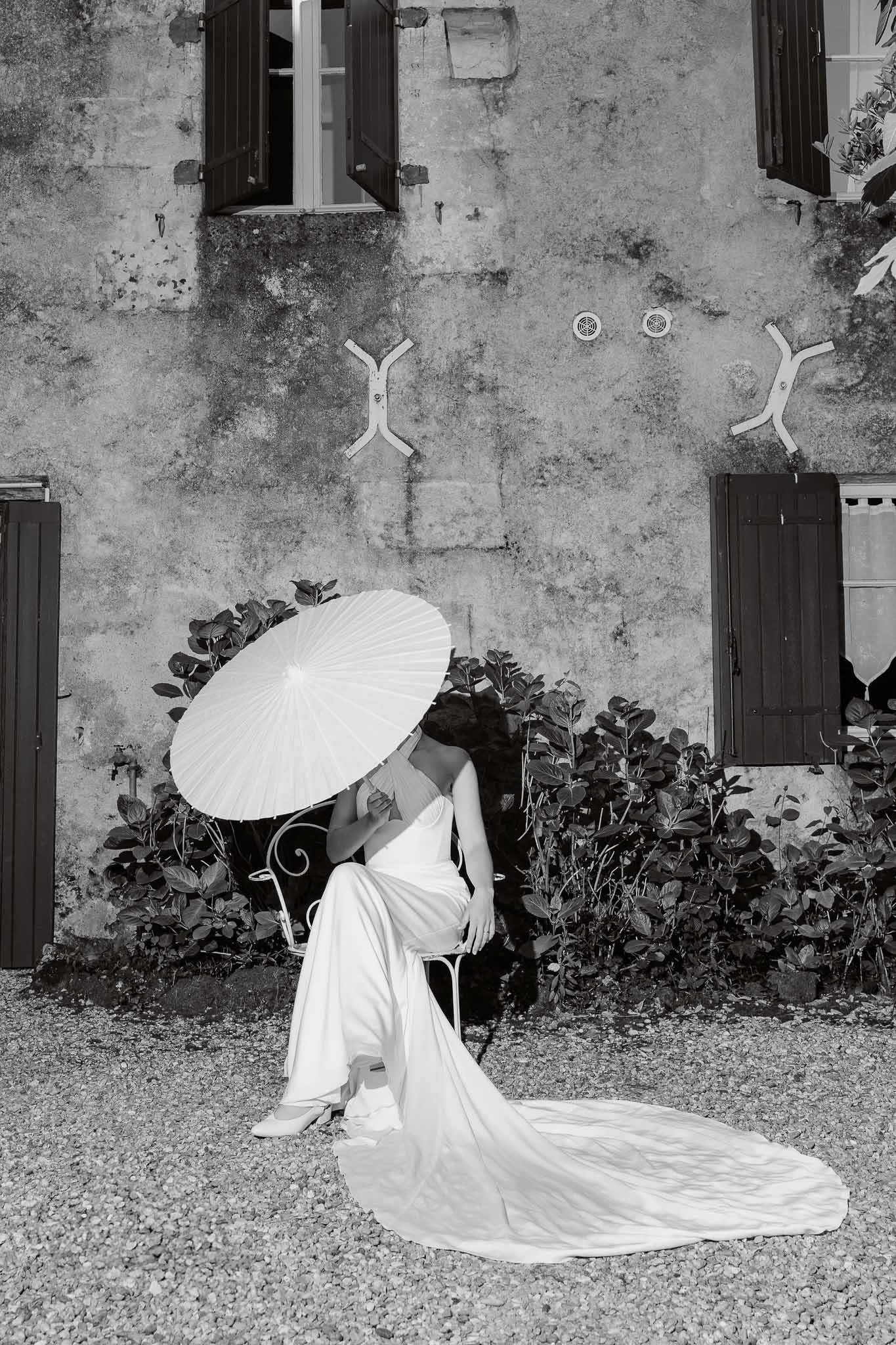 Black and white bride seated on wrought-iron chair with paper parasol hiding face and strapless gown train on gravel