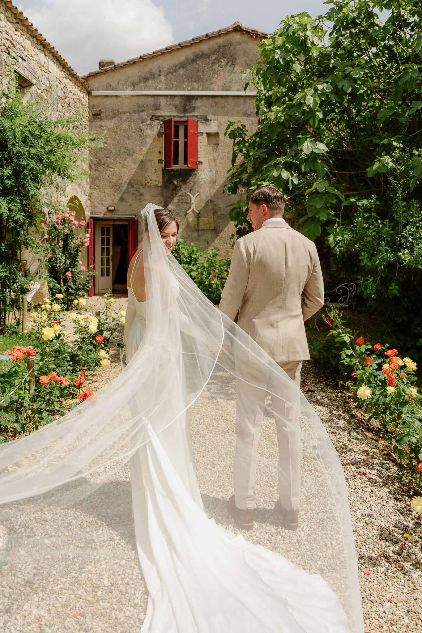 Couple walking through rose-lined courtyard from behind with cathedral veil fanning across gravel before red-shuttered far...