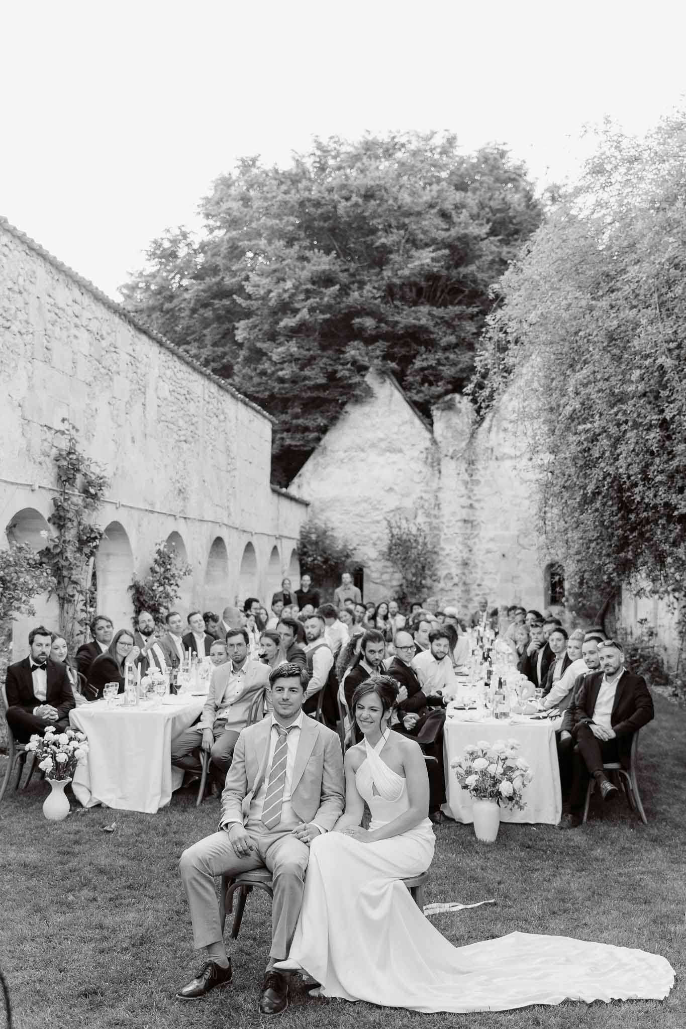 Black and white couple seated before sixty guests at courtyard reception with stone arcades and climbing vines
