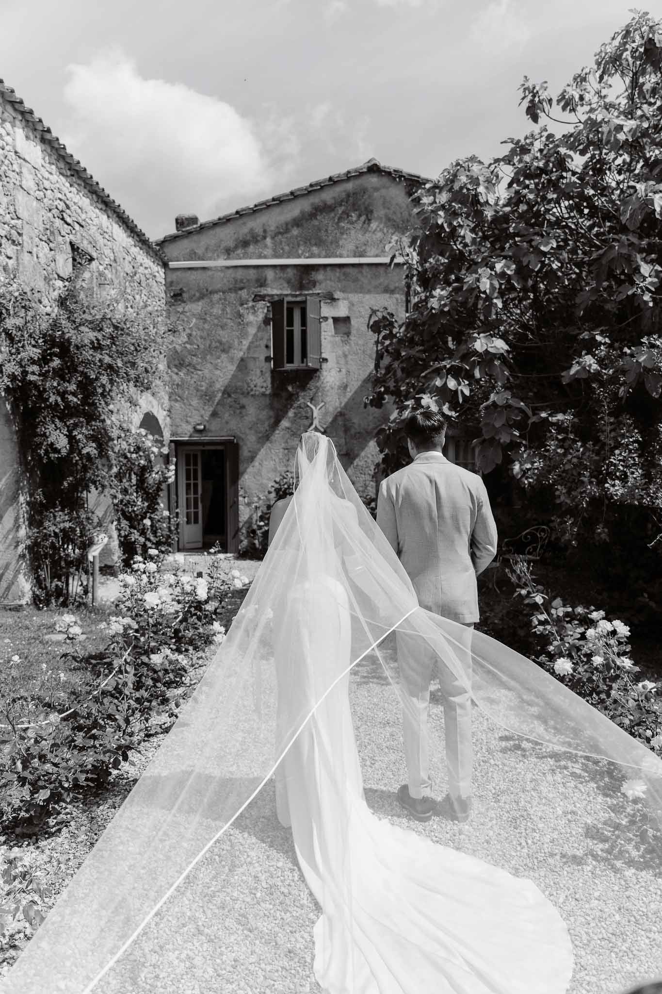 Black and white couple walking away with cathedral veil trailing across gravel toward vine-covered stone building