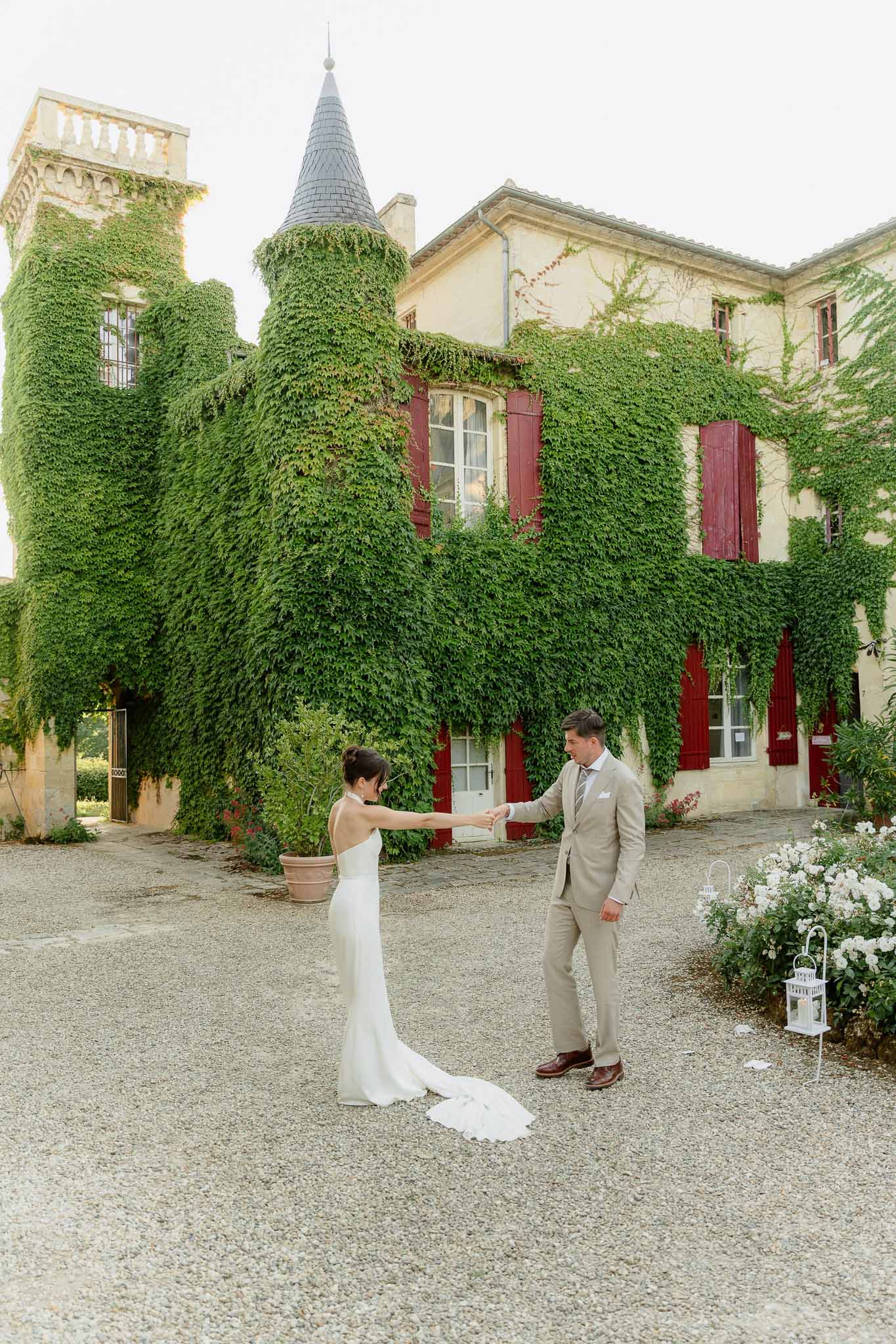 Groom taking bride's hand on gravel before ivy-covered chateau with turret and burgundy shutters beside white lanterns