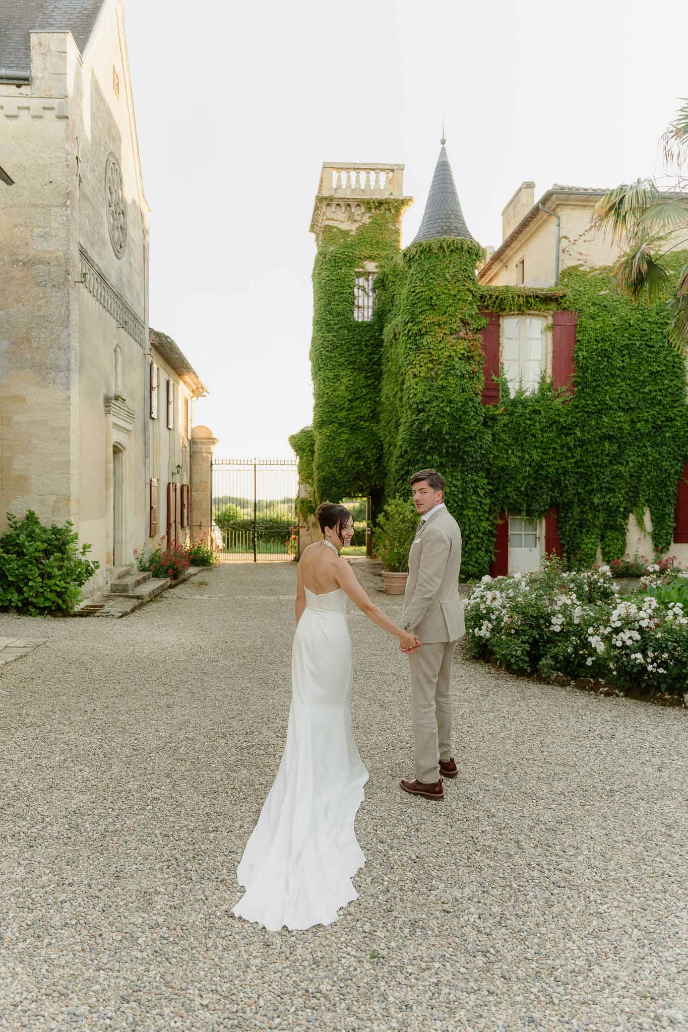 Couple holding hands on chateau gravel courtyard with ivy turrets and wrought iron gates opening to countryside