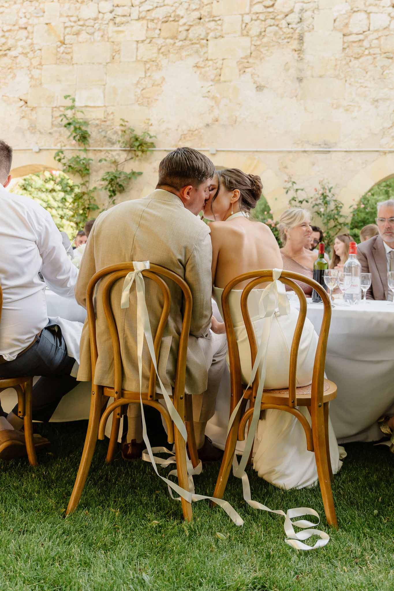Couple kissing at sweetheart table from behind with ribbon-tied chairs and guests at surrounding tables