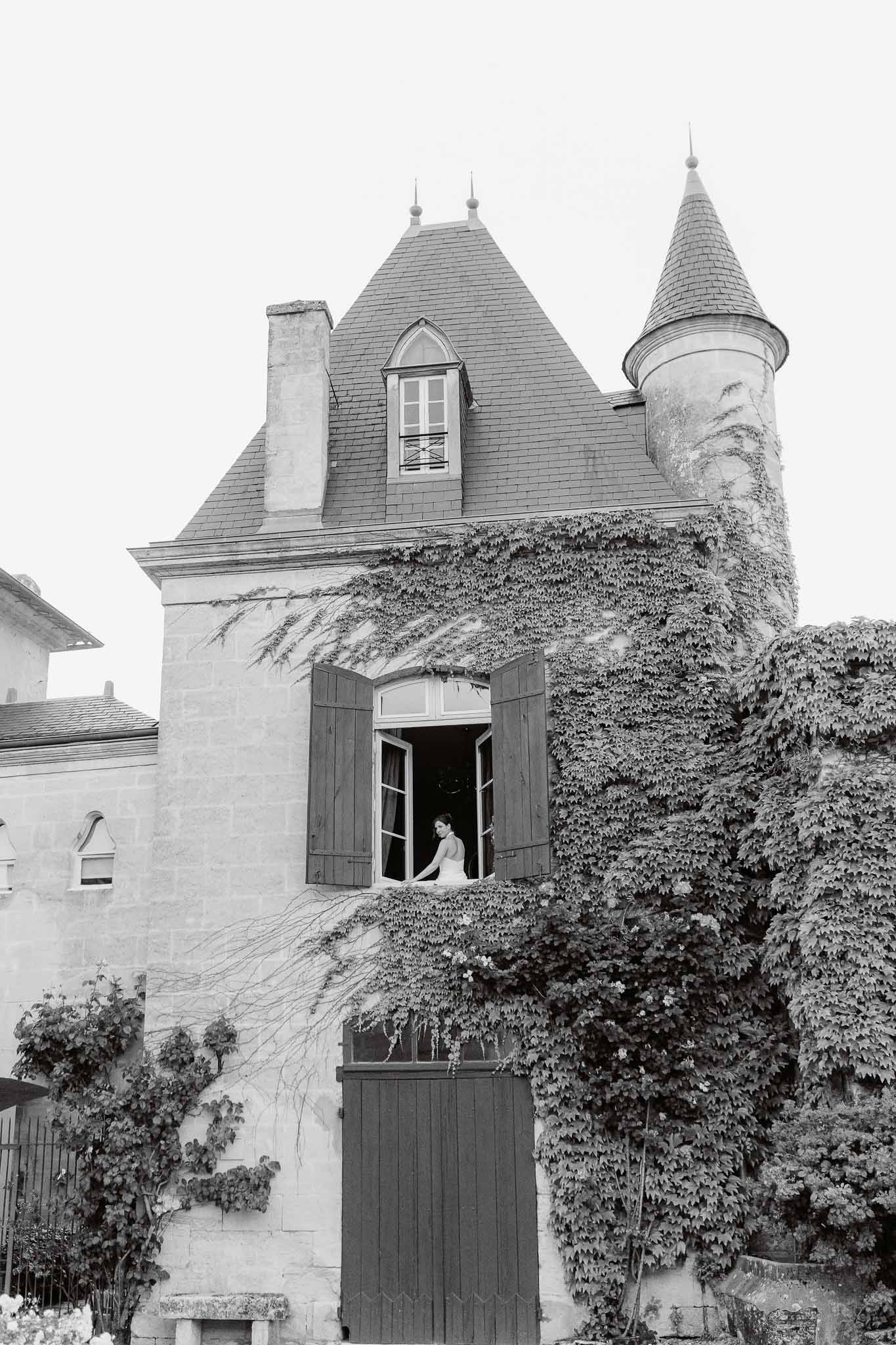 Black-and-white shot of bride at open chateau window with ivy-covered stone facade and slate turret roof