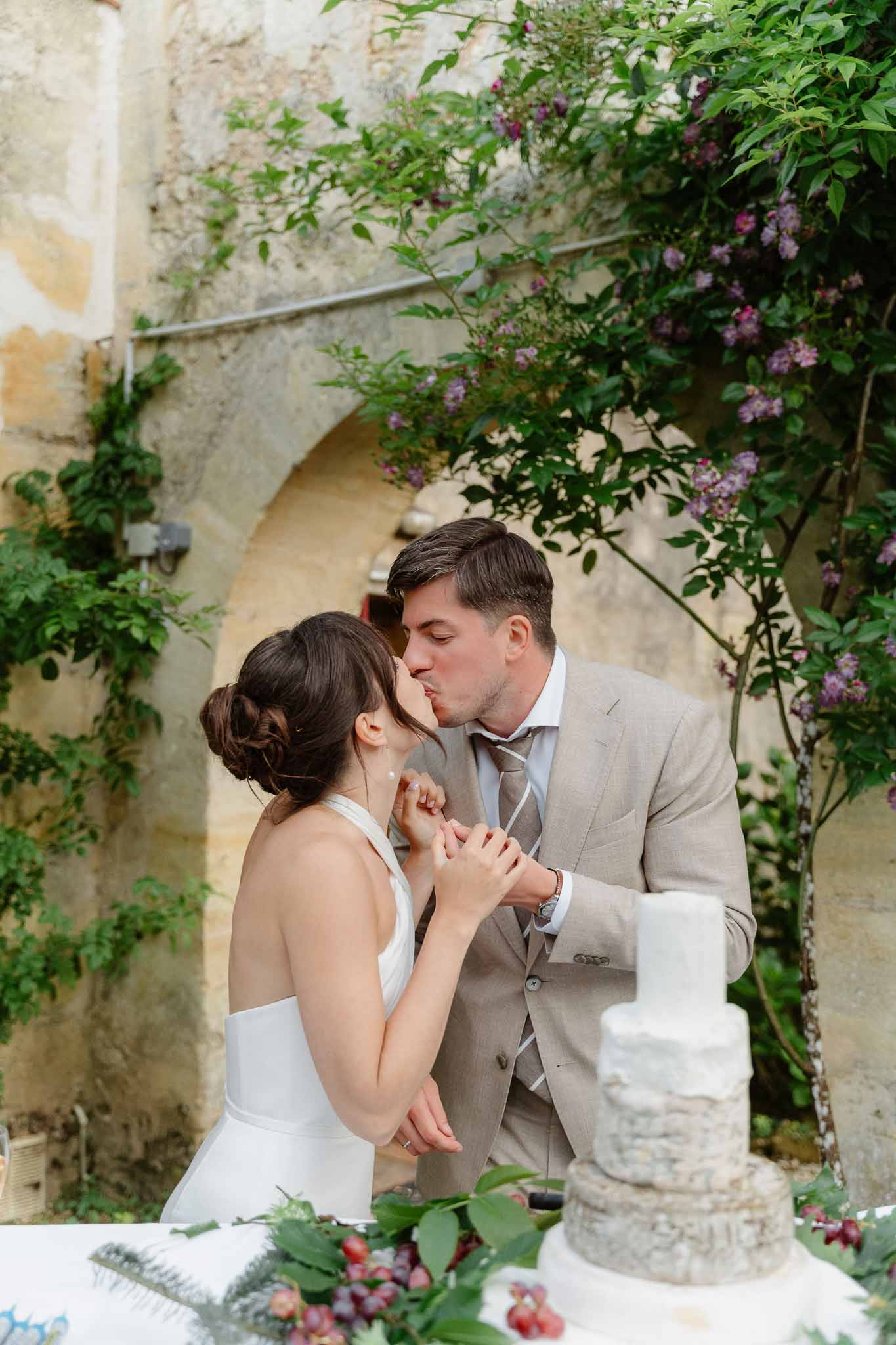Bride and groom kiss during cake cutting under a stone archway with climbing vines at outdoor reception