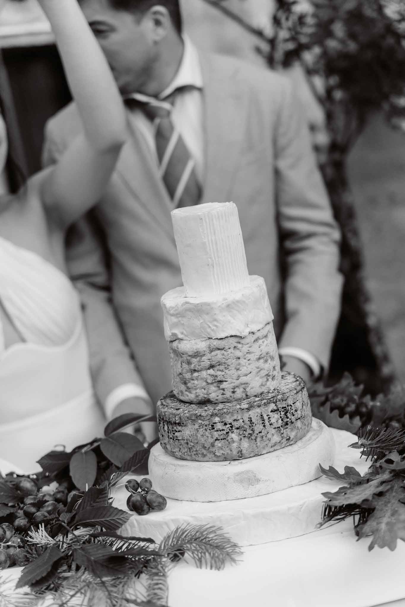 Black and white close-up of couple cutting cheese wheel tower decorated with foliage and berries