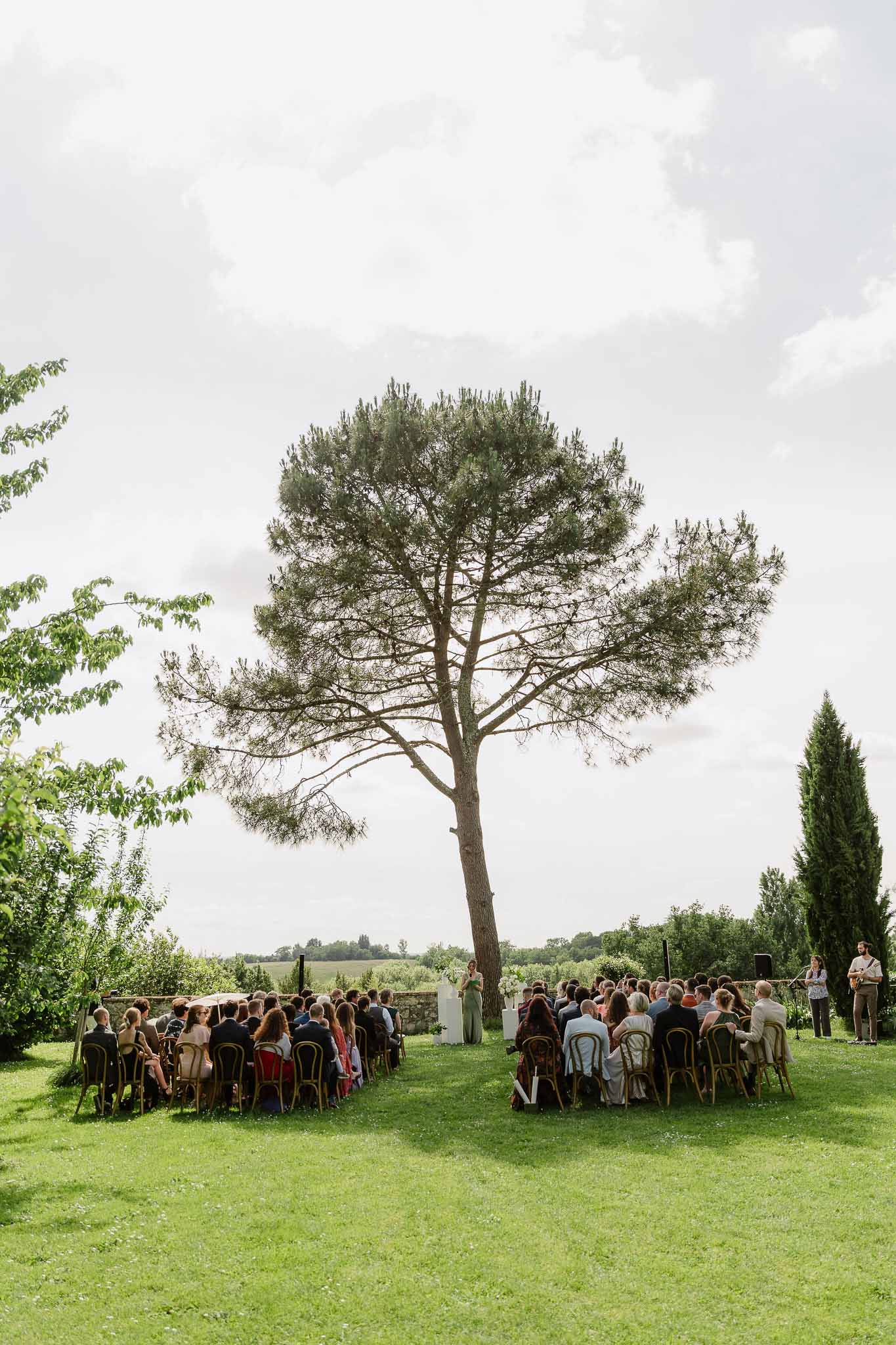Ceremony under umbrella pine on lawn with bistro chairs, acoustic guitarist, and countryside beyond stone wall