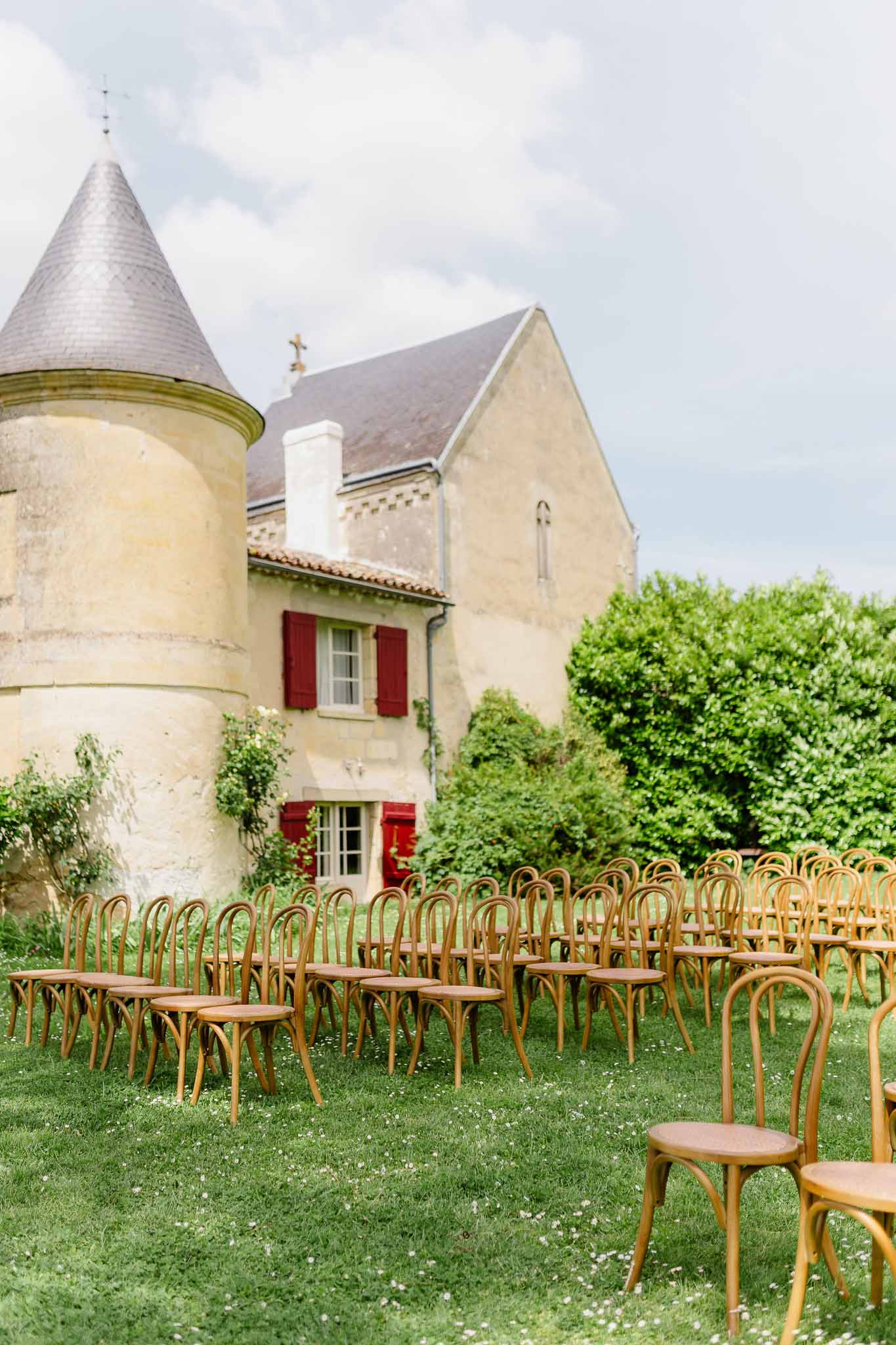 70 honey-toned bentwood chairs in semicircle on lawn before cream chateau with conical turret and red shutters