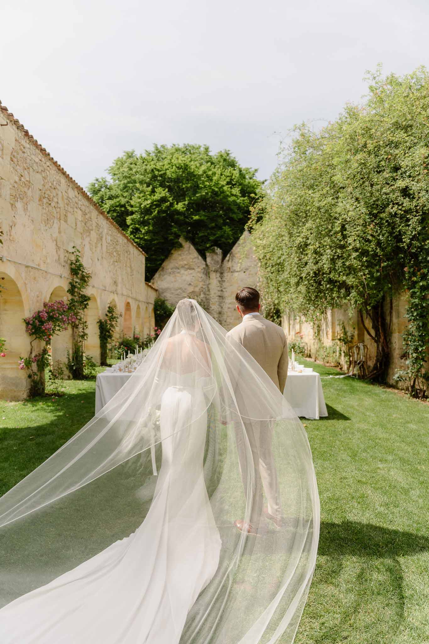 Bride and groom walking toward outdoor reception table, cathedral veil billowing across lawn at limestone chateau