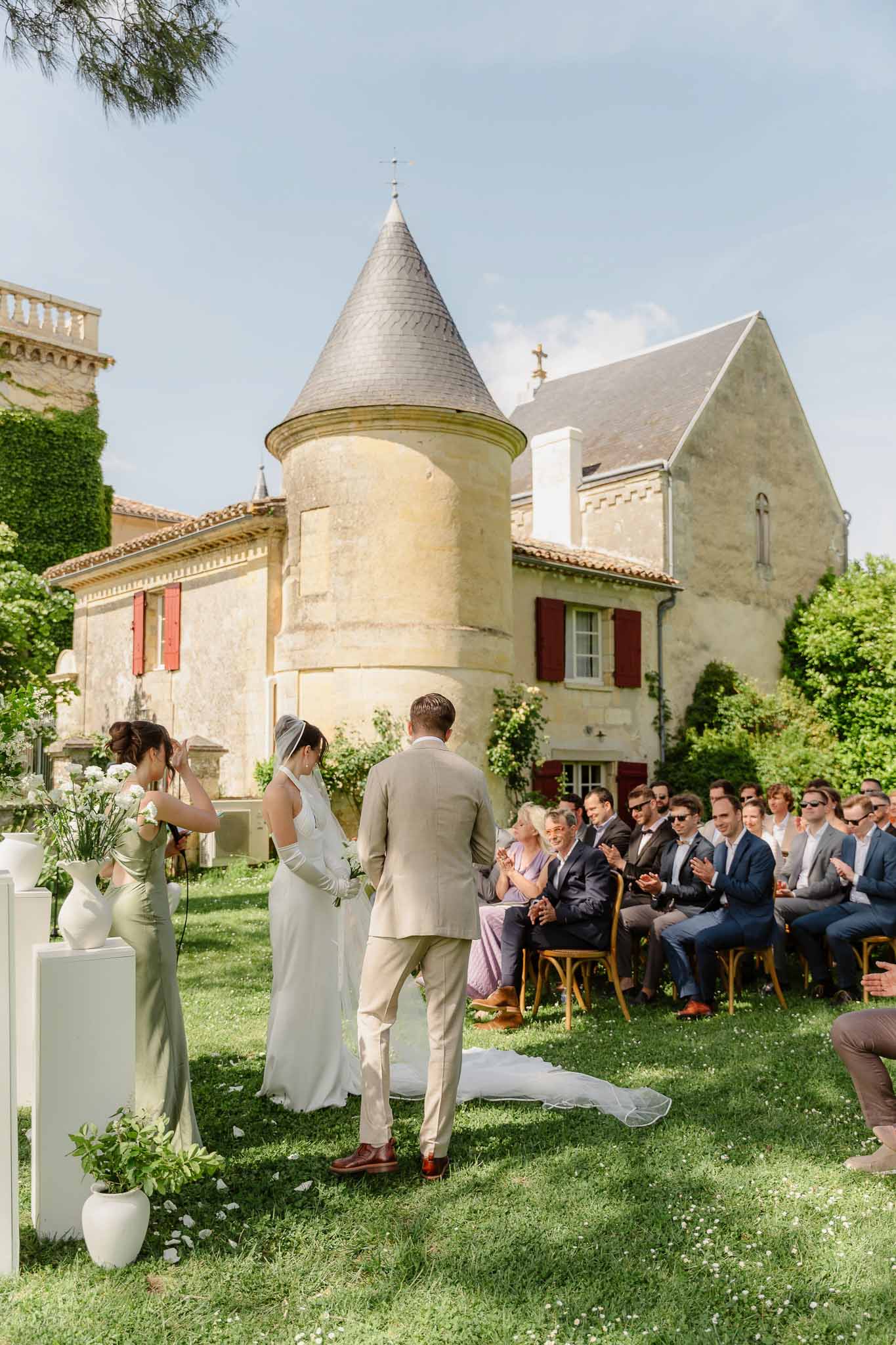 Couple holding hands at altar before stone tower with white pedestals and sage-dressed bridesmaid