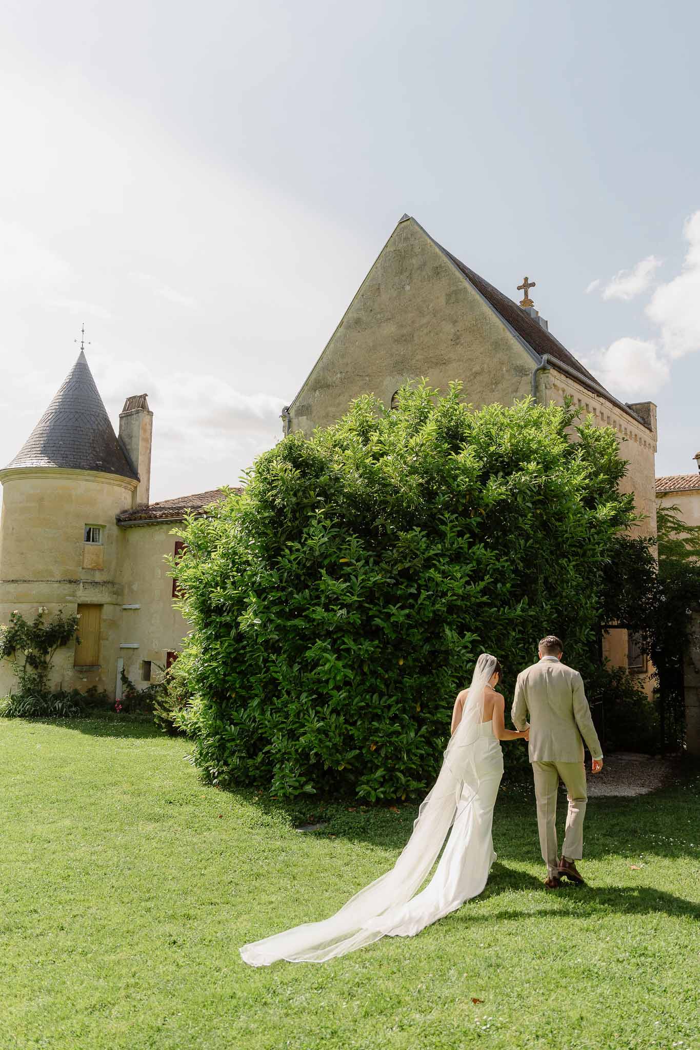 Bride and groom walking across lawn toward chateau tower and chapel with cathedral veil trailing on grass