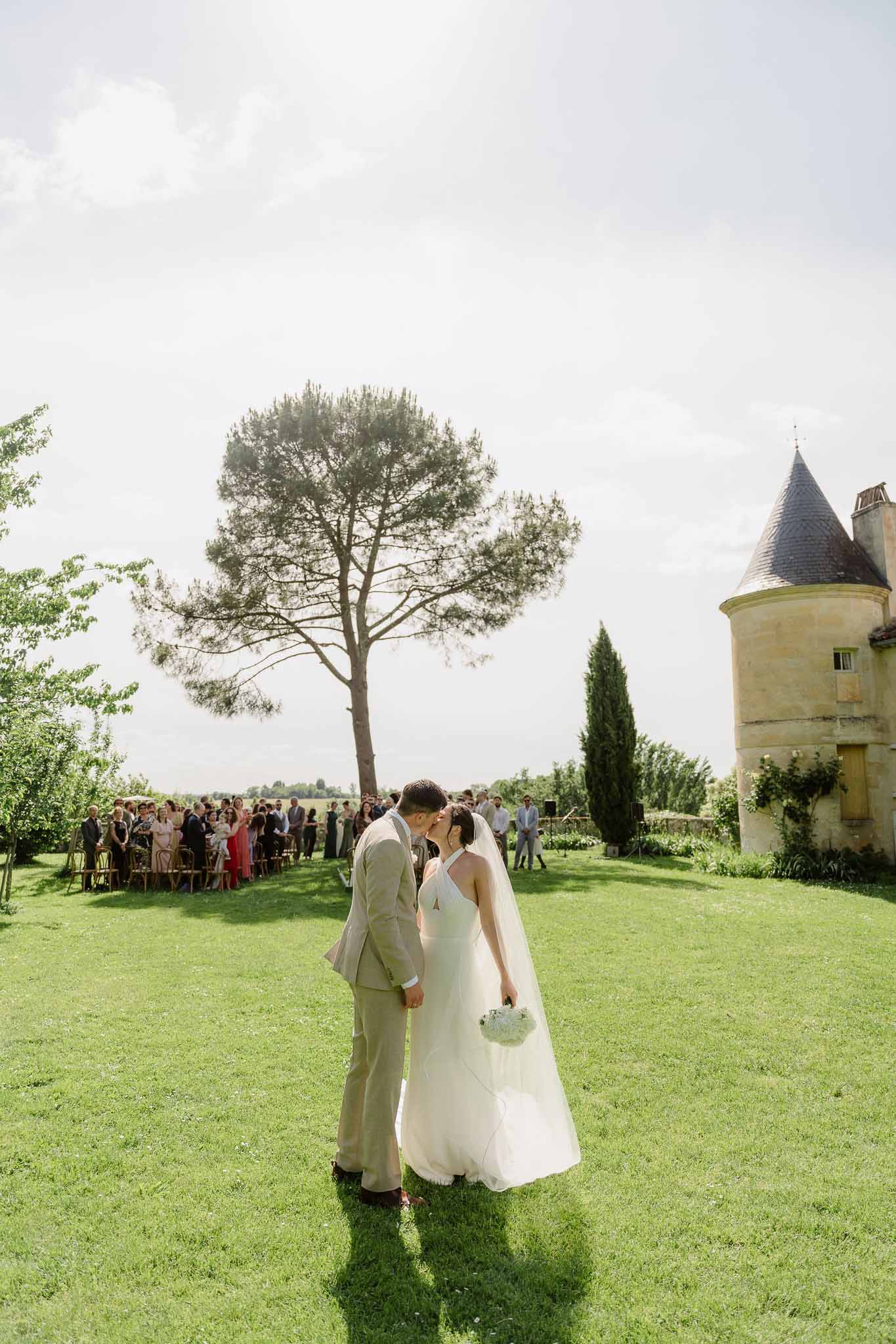 Outdoor ceremony first kiss on chateau lawn with seated guests and stone tower backdrop