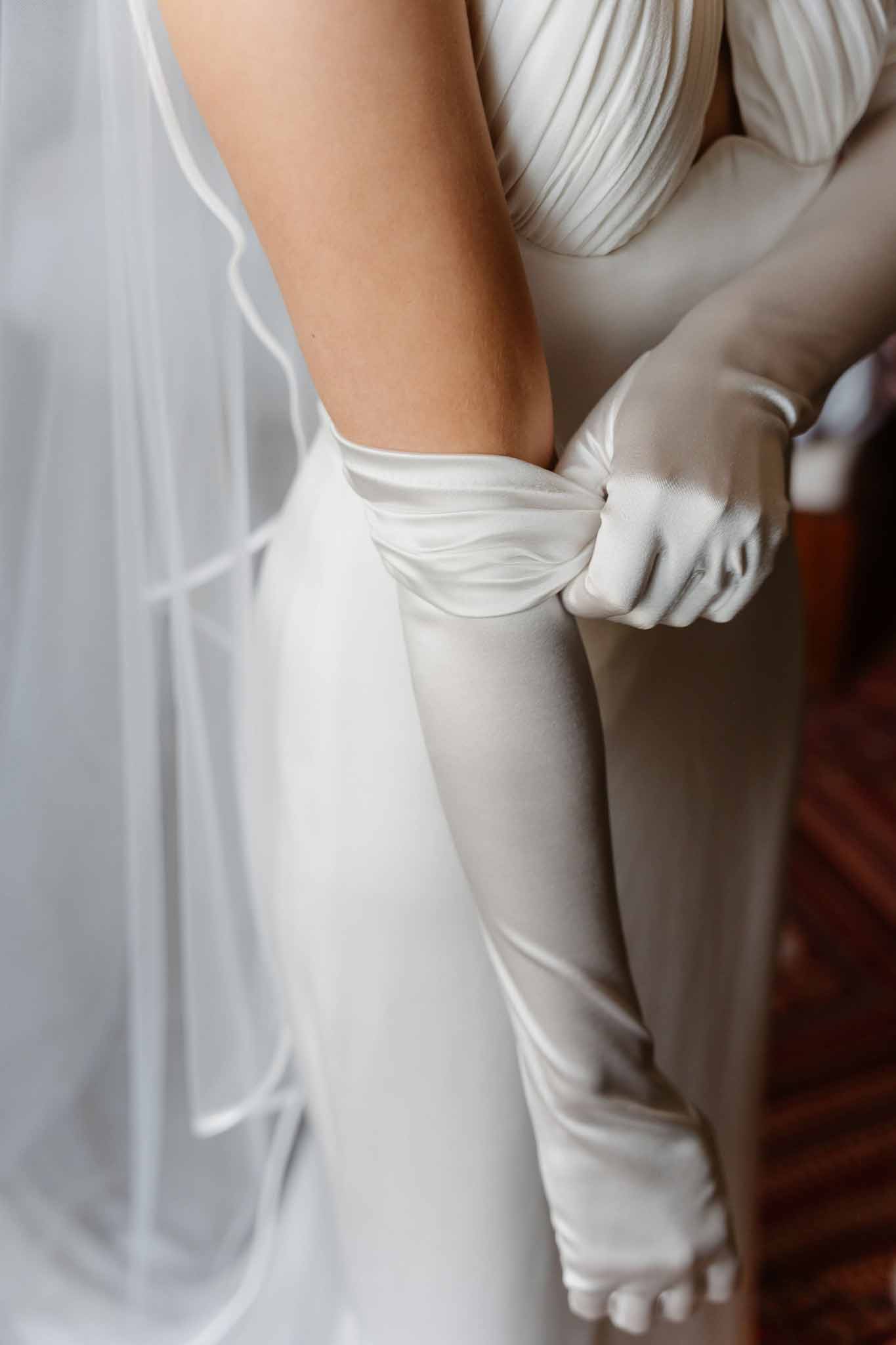Close-up of bride pulling on ivory satin opera-length glove over pleated gown