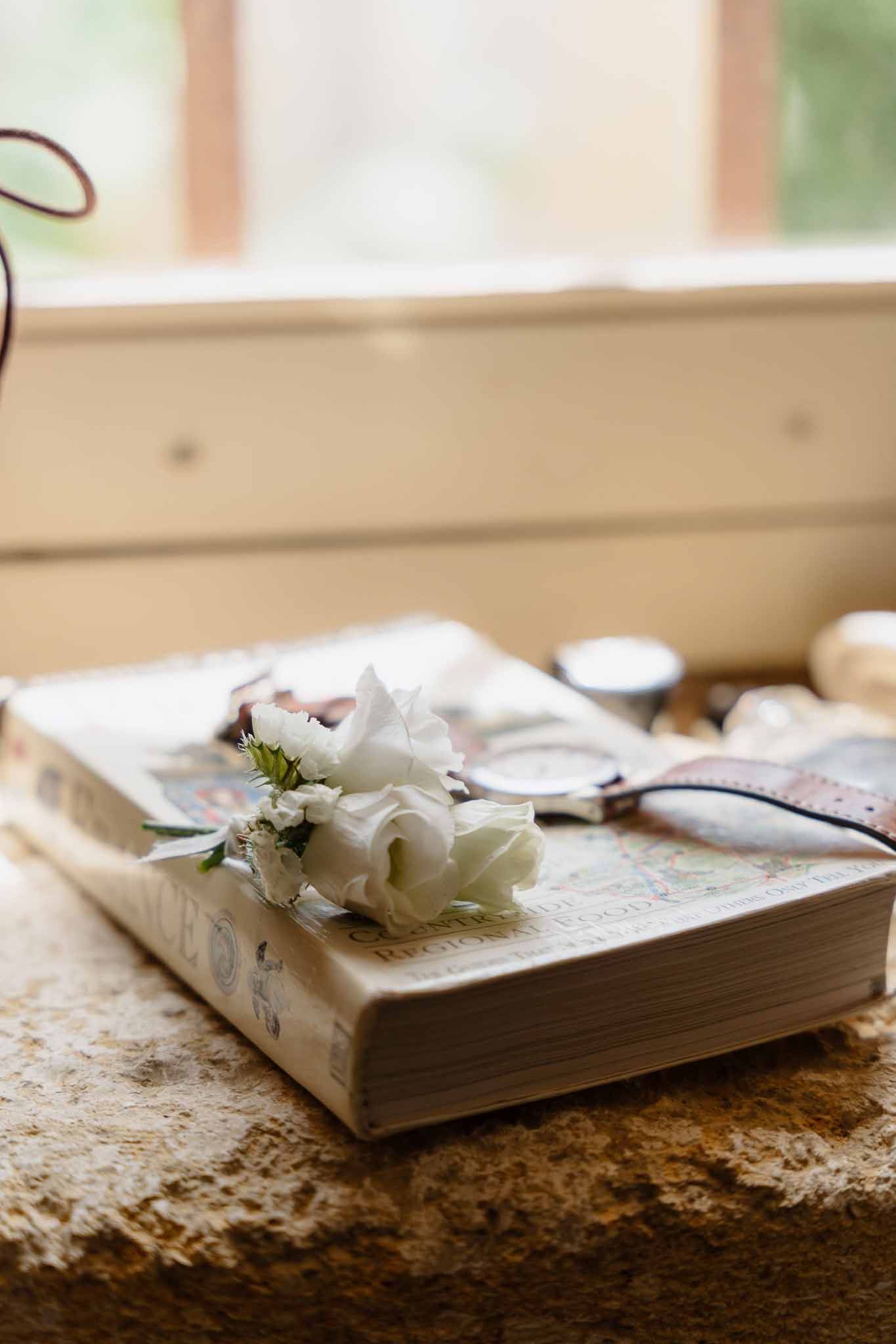 White lisianthus boutonniere on hardcover book beside watch and cufflinks on stone windowsill