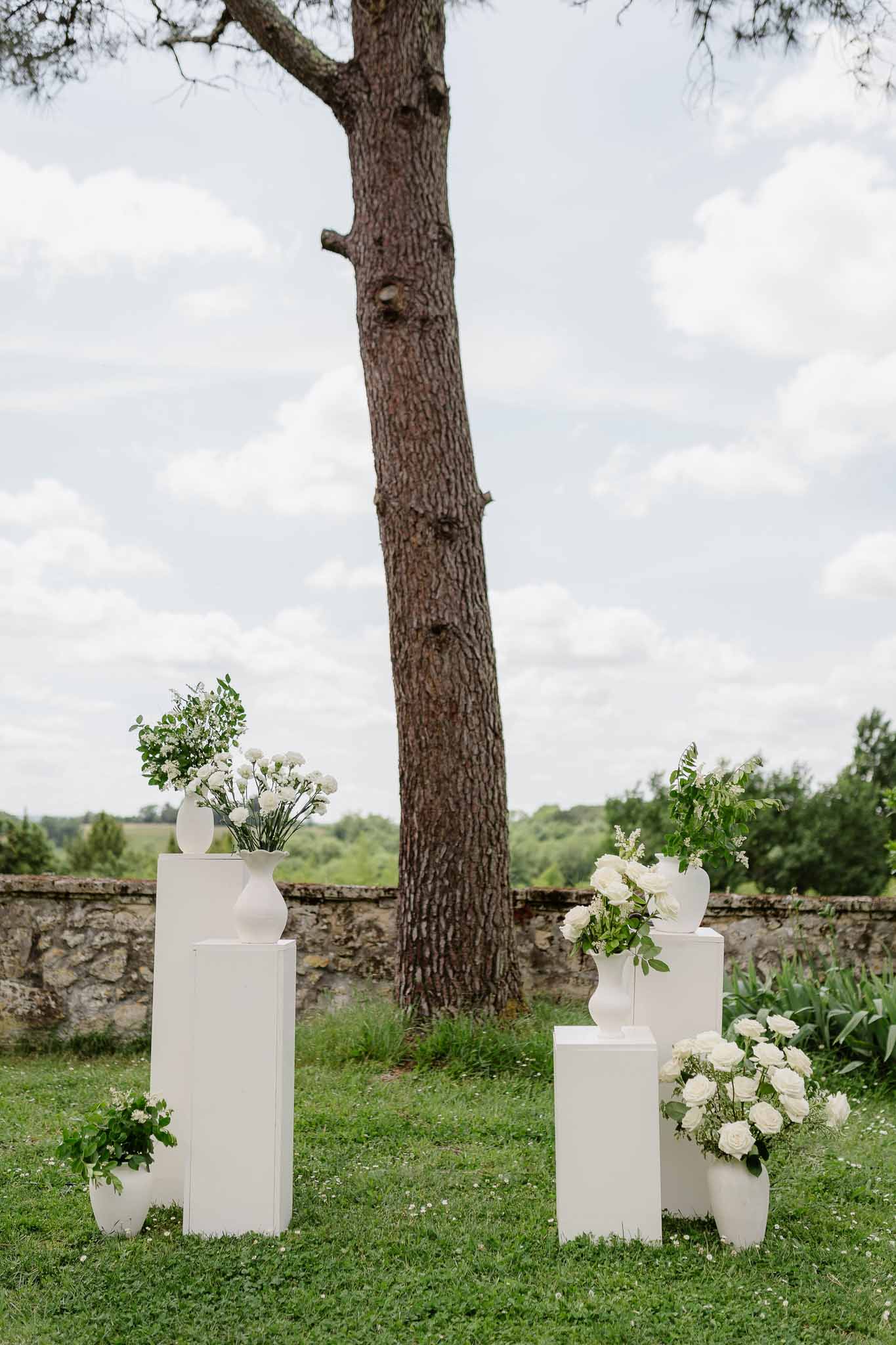 White pedestals with white rose and ranunculus arrangements on a lawn with a stone wall behind