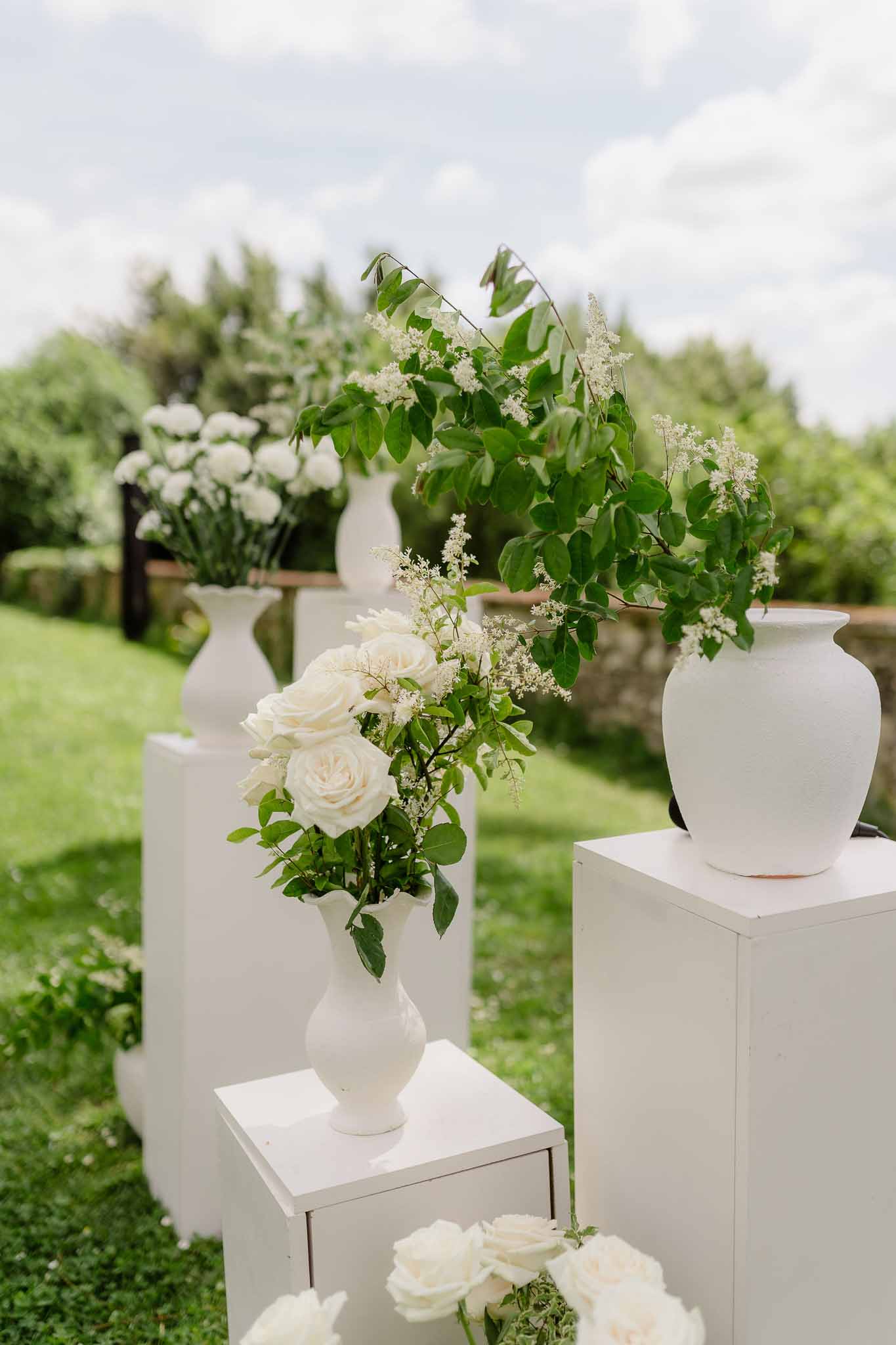 White ceramic vases on plinths with ivory roses and green foliage ceremony aisle arrangements