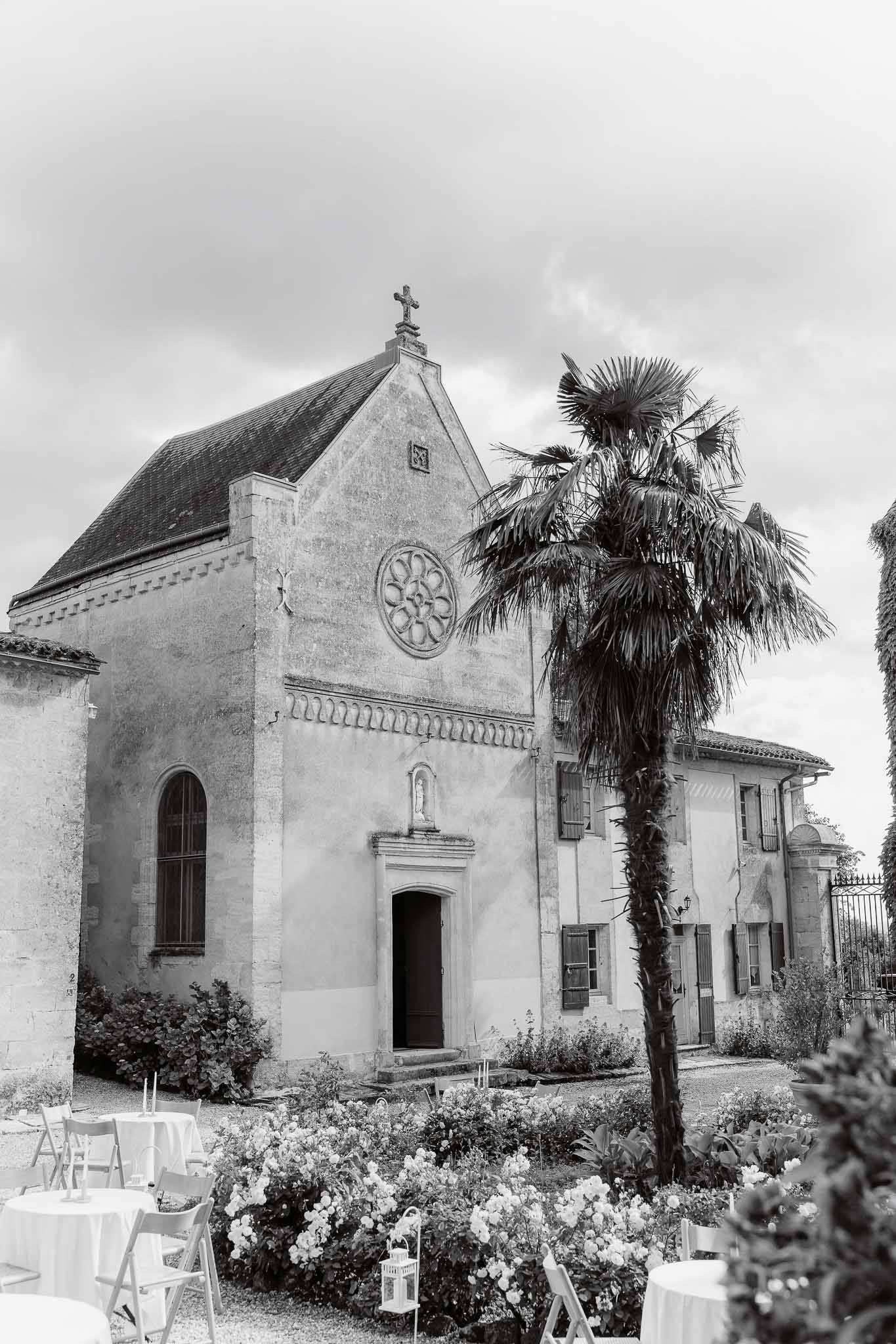 Romanesque estate chapel with rose window and palm tree beside cocktail tables on garden in B&W