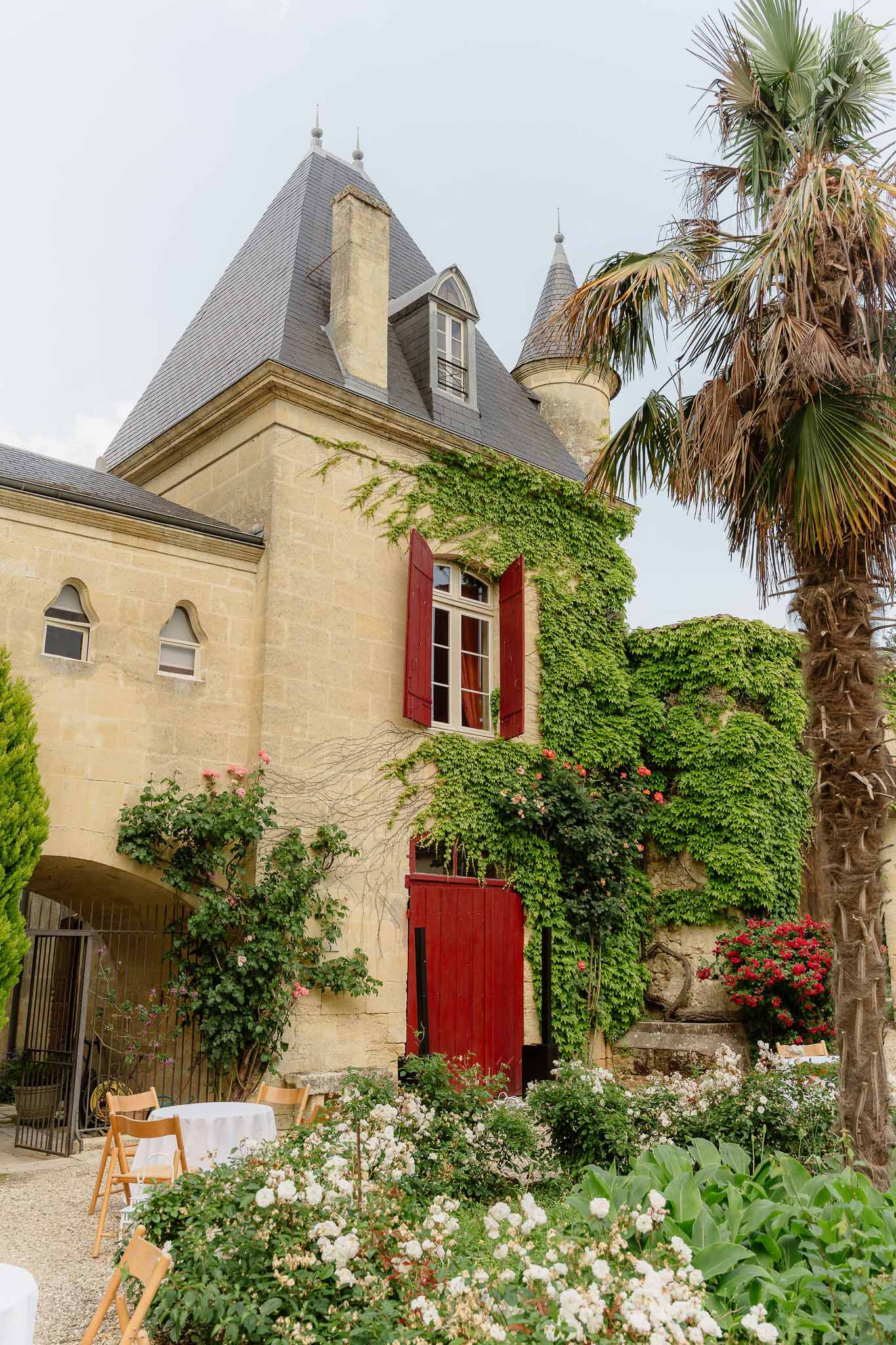 Honey-stone chateau with slate turrets, red shutters, and ivy beside white-clothed courtyard tables