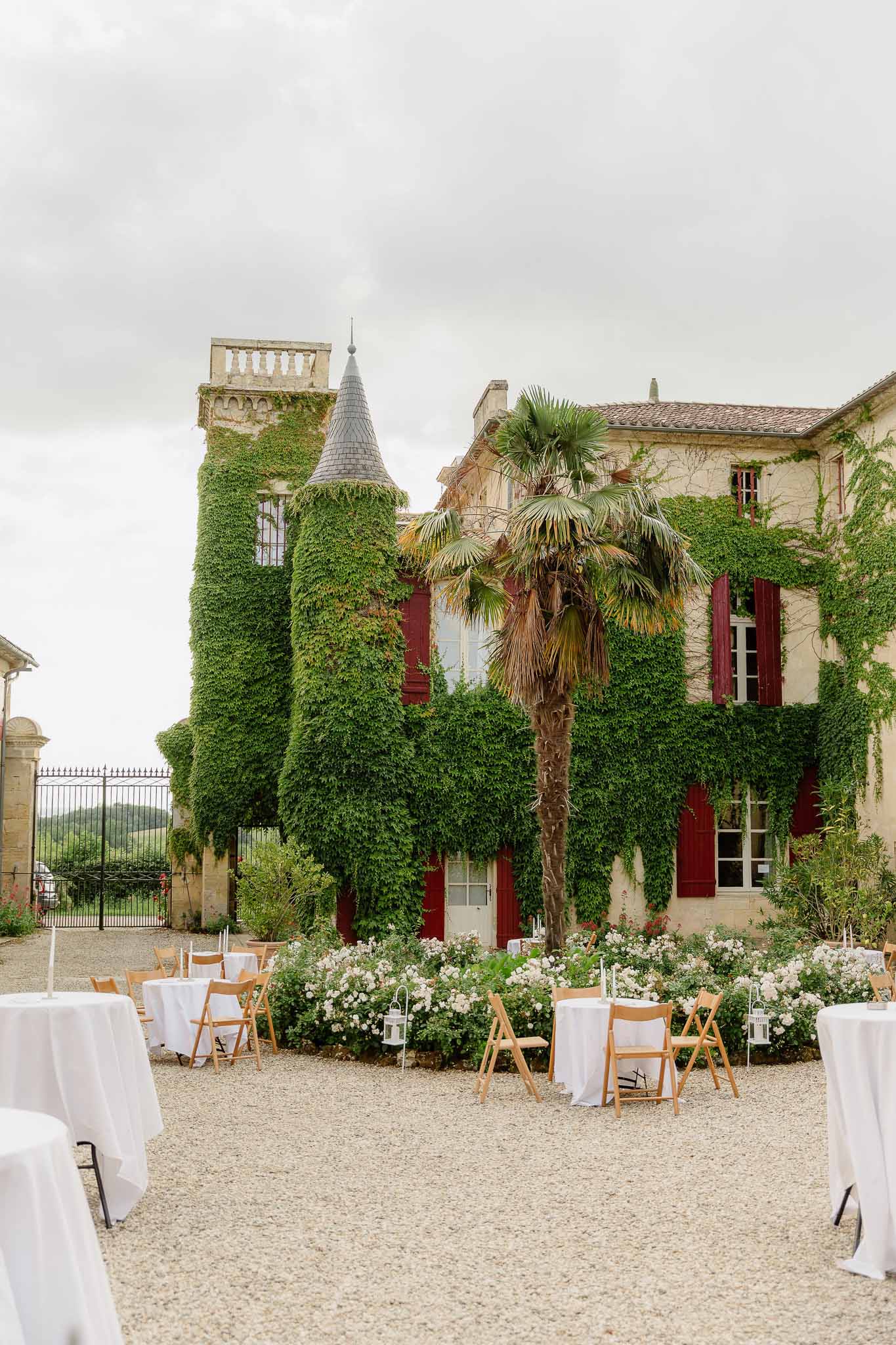 Round white tables and wood chairs on gravel courtyard before ivy-covered chateau with conical tower