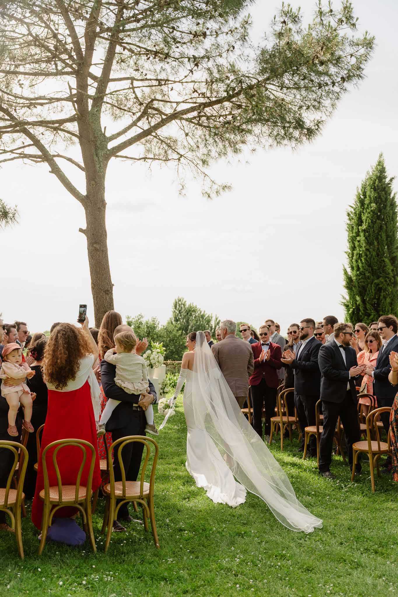 Bride with opera gloves and cathedral veil walking down outdoor aisle with standing guests applauding