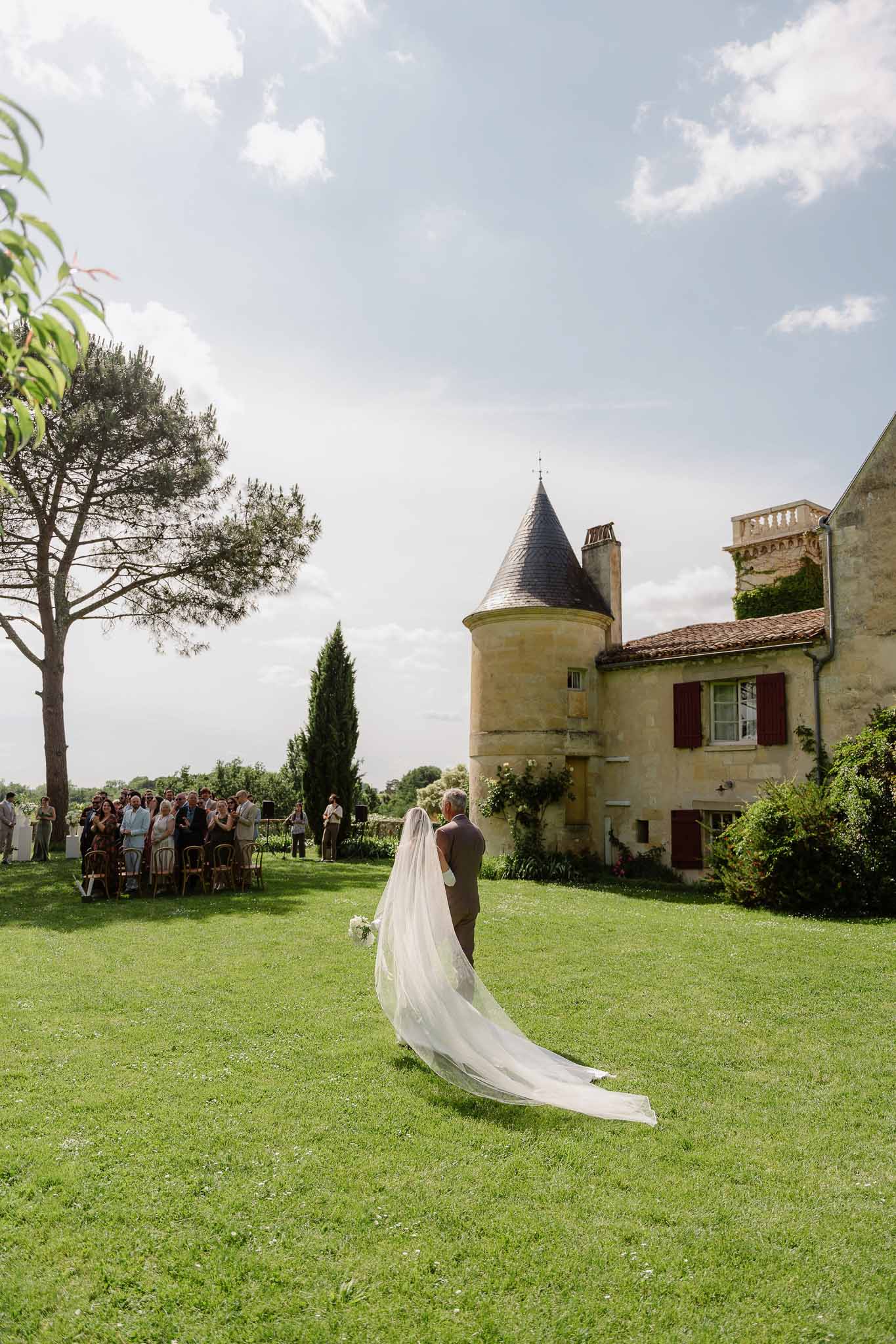 Bride with cathedral veil escorted across lawn toward 40 seated guests with turreted chateau behind