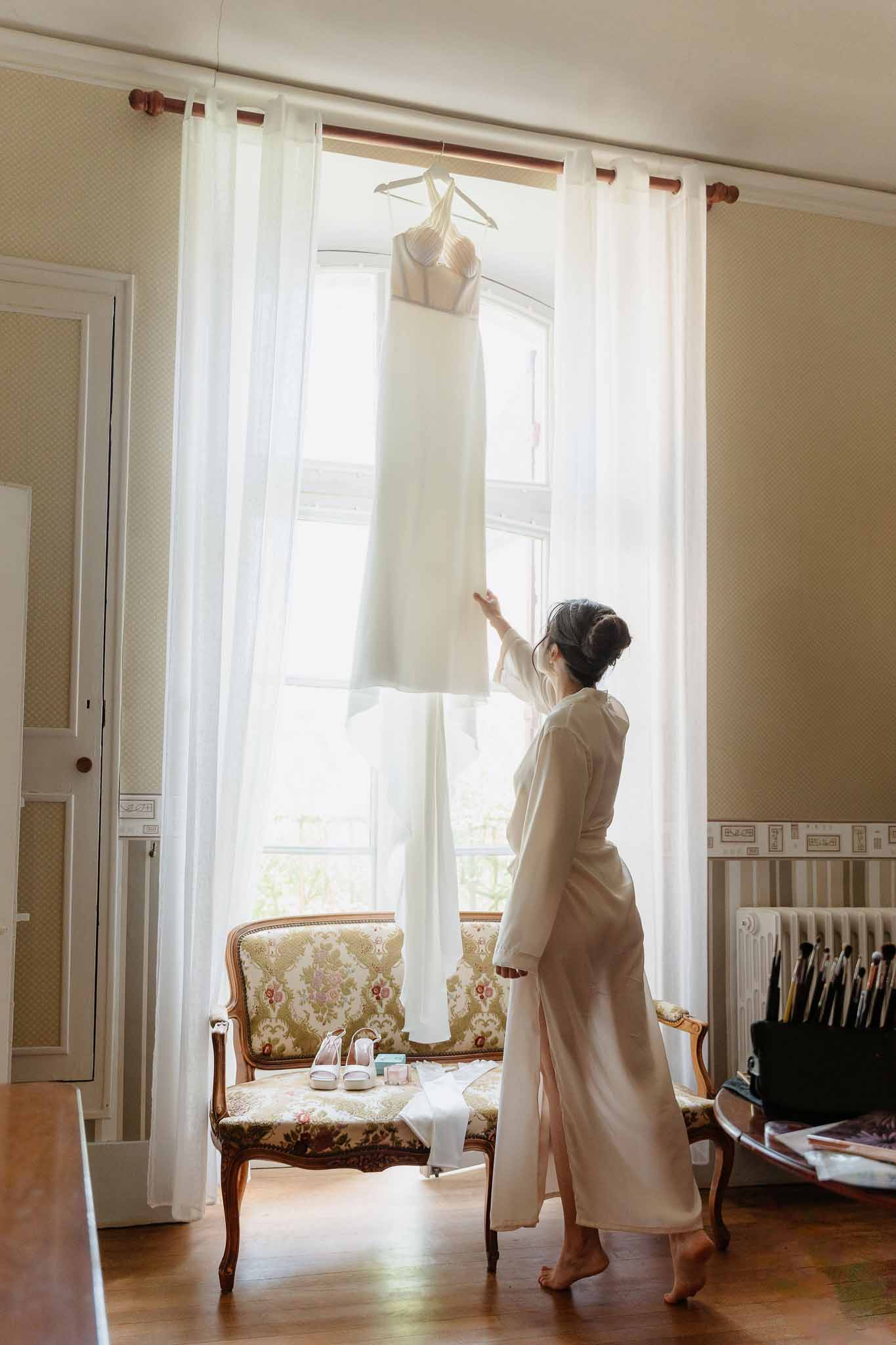 Bride in ivory robe reaching for column gown hanging by window with heels on floral settee