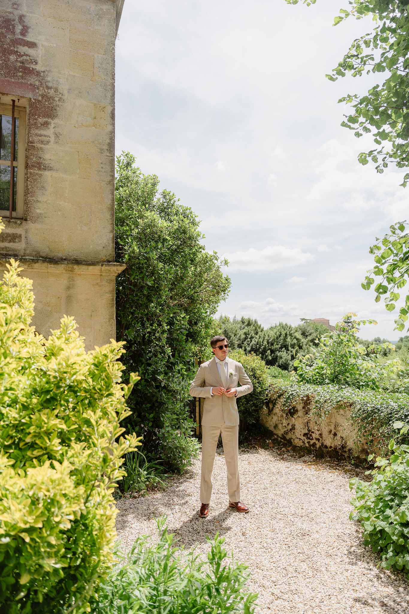 Groom adjusting sand suit button with sunglasses on gravel path beside chateau hedges