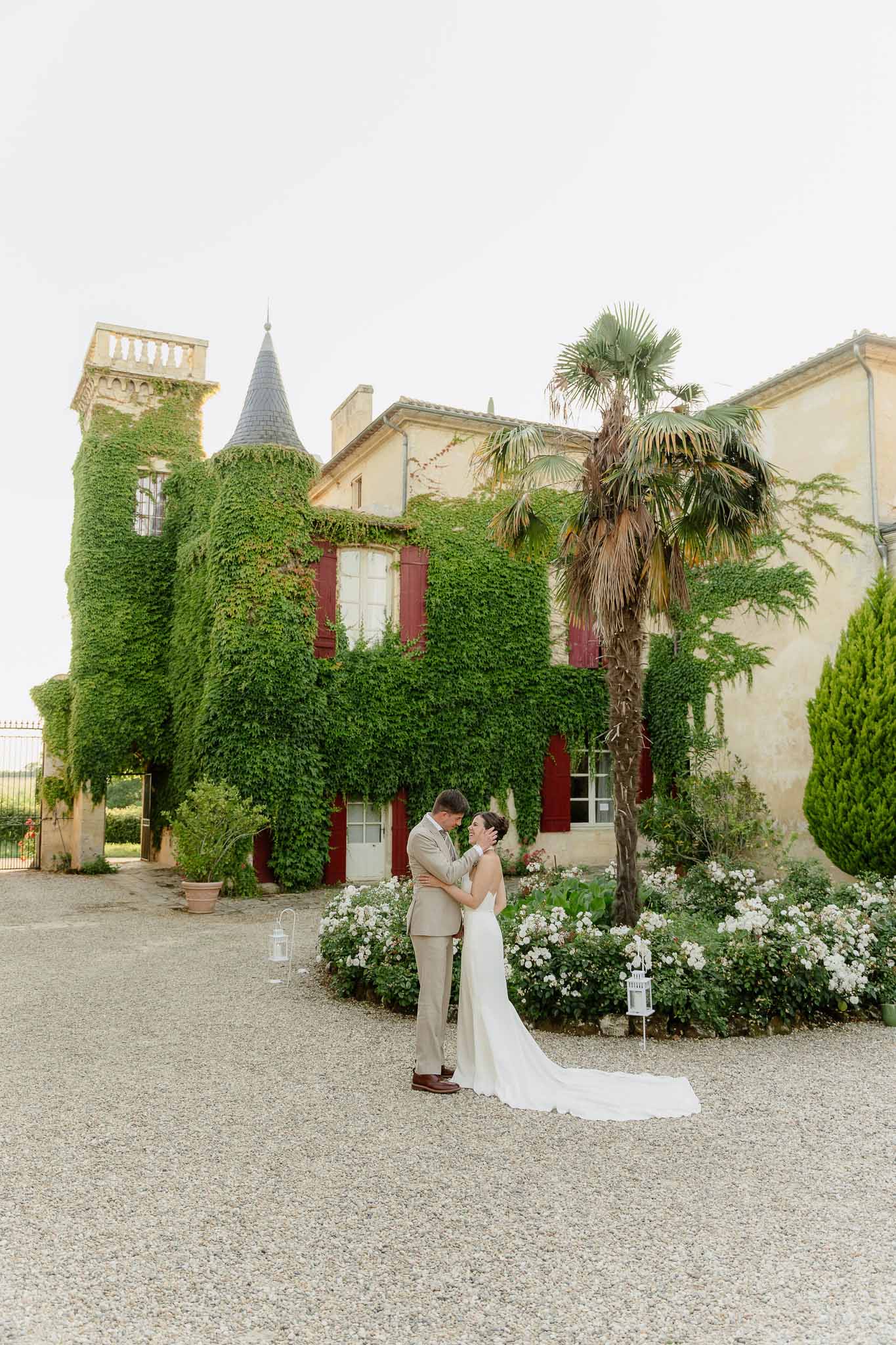 Couple touching foreheads on gravel drive before ivy-covered chateau with conical tower and red shutters