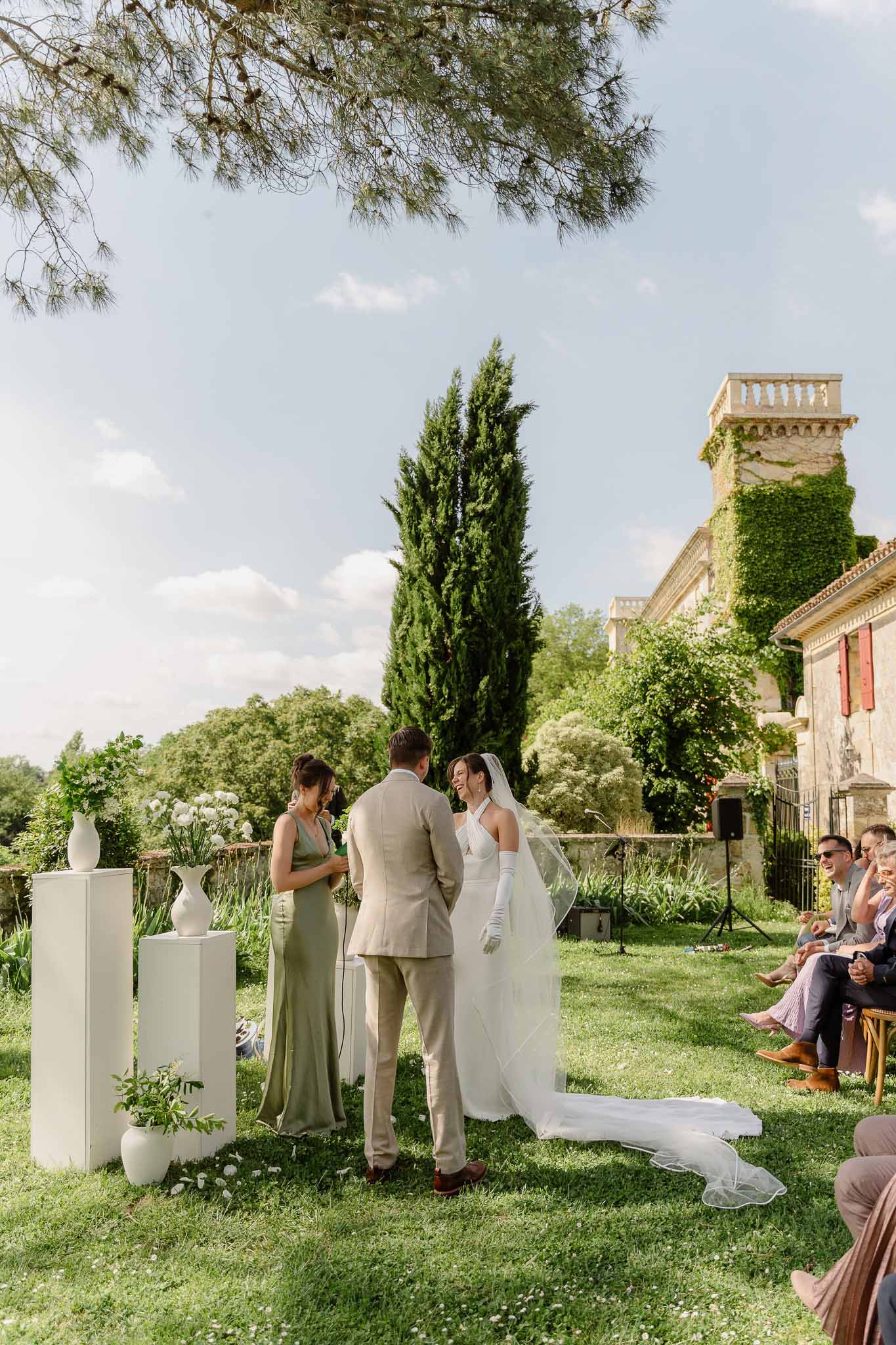 Outdoor ceremony on lawn before ivy-covered chateau with white allium arrangements on plinths as altar backdrop