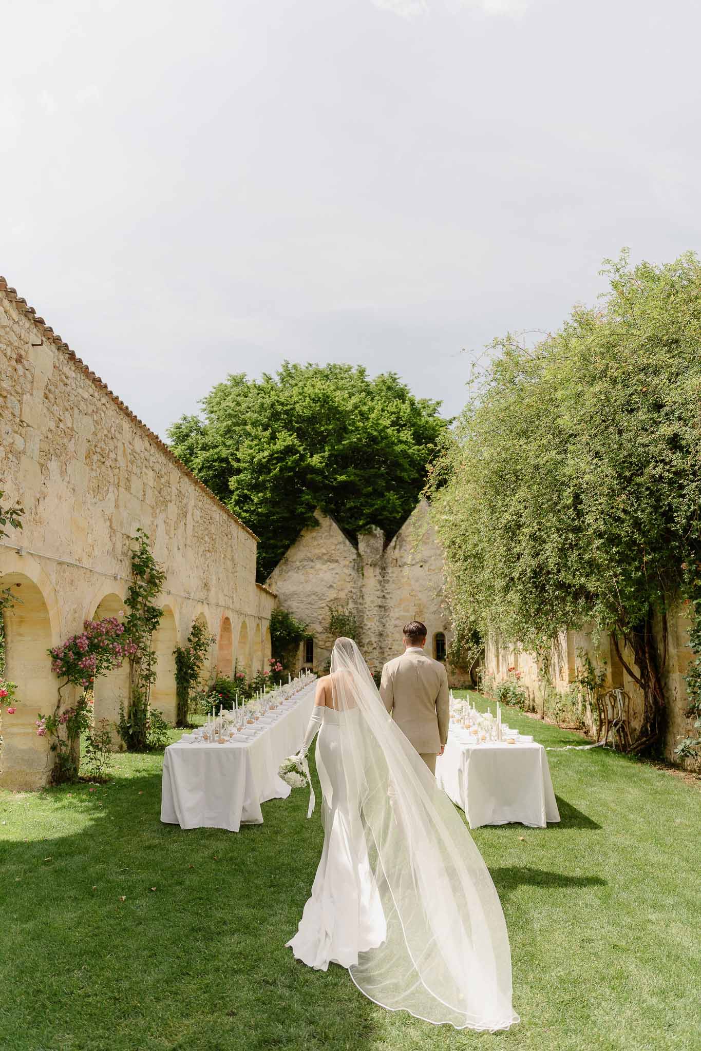 Bride and groom walking between long banquet tables in limestone courtyard with cathedral veil trailing on lawn
