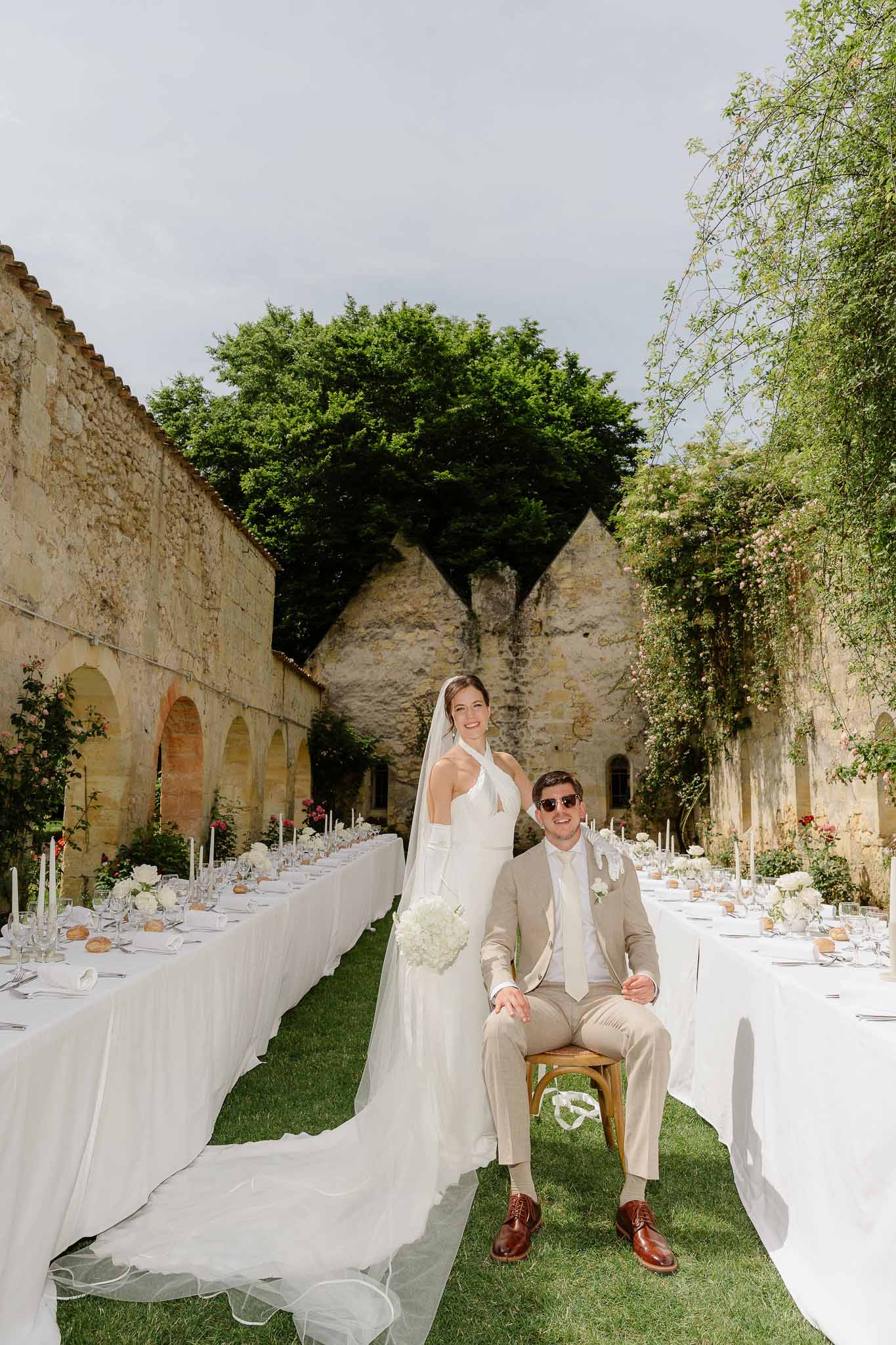 Bride and groom posing between two parallel banquet tables set for outdoor reception in stone courtyard of French abbey