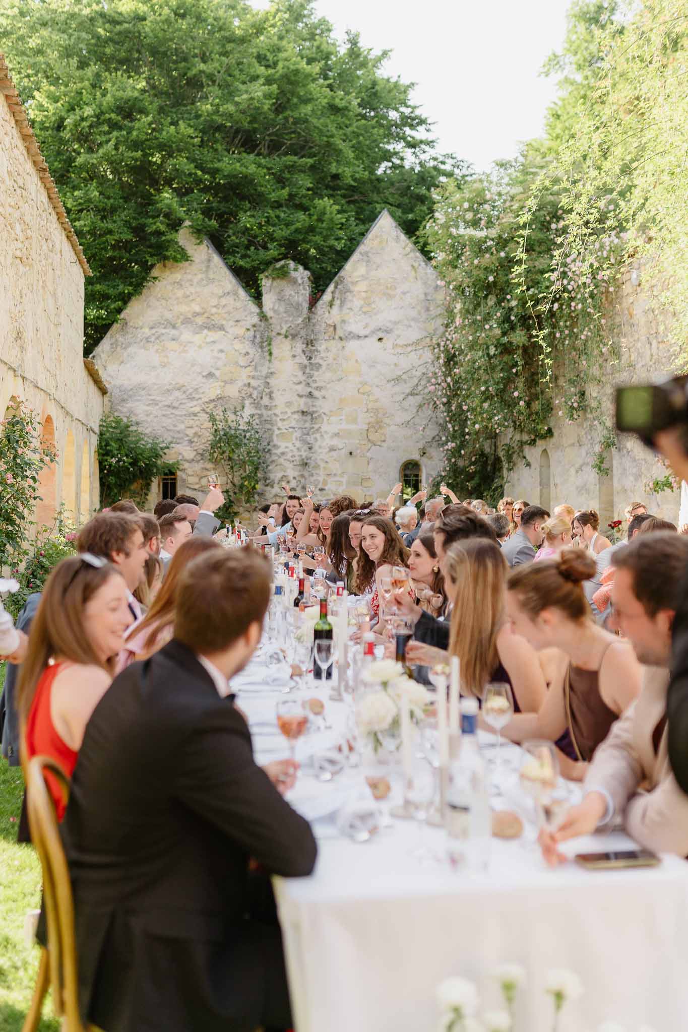 Forty guests at long banquet table in walled courtyard with white ranunculus centerpieces and climbing roses