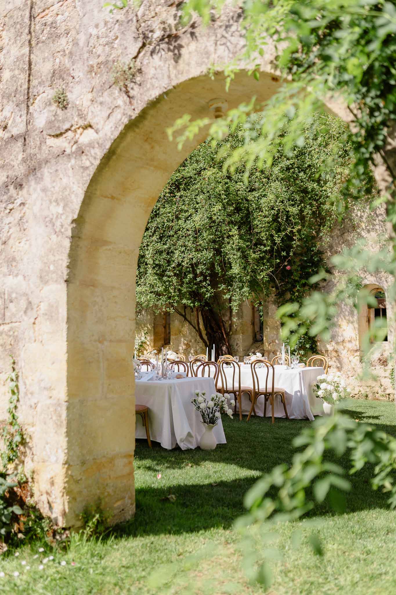 Outdoor reception tables with white linen and rattan chairs framed through stone archway with foliage