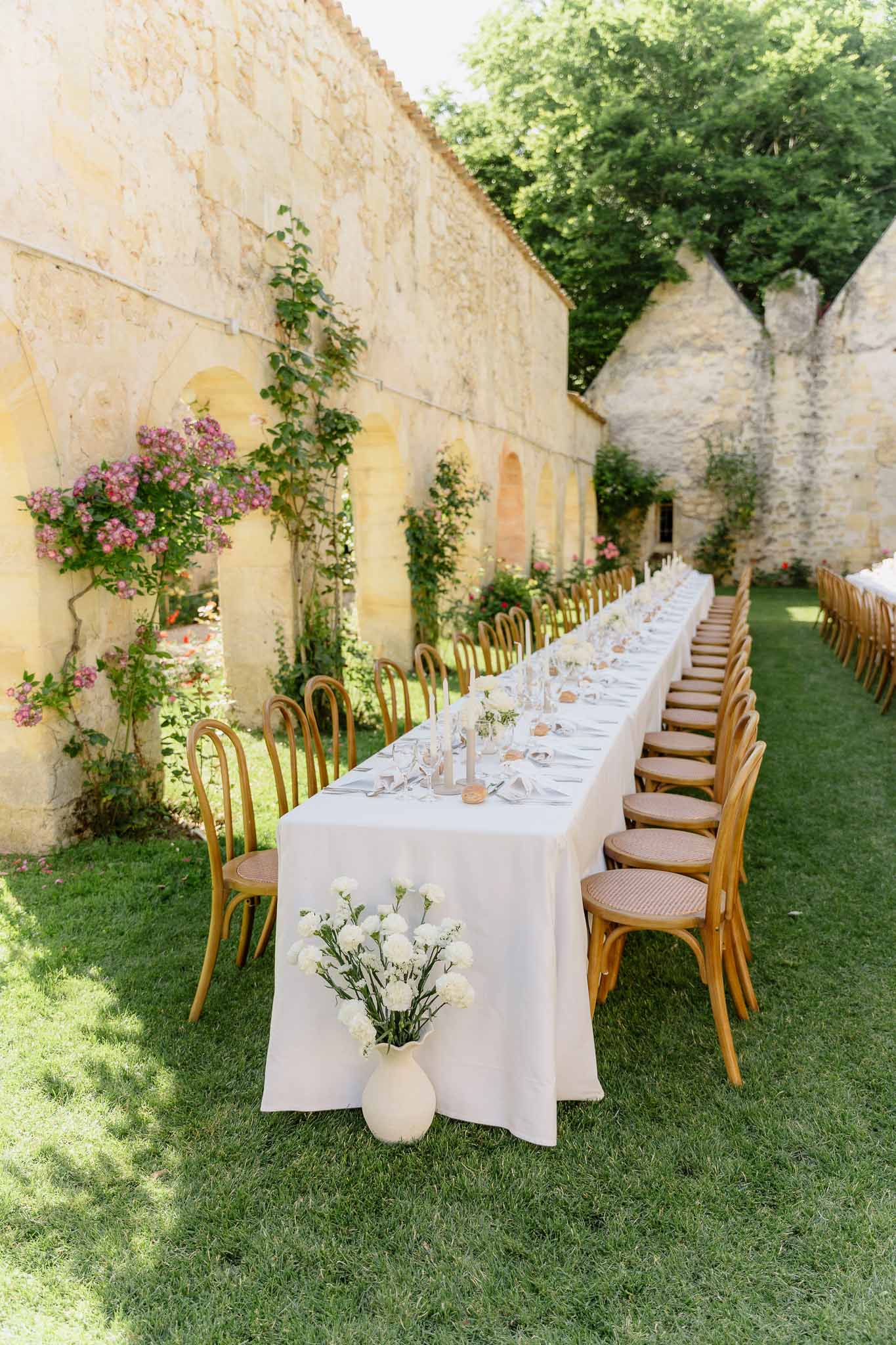 Long table with bentwood bistro chairs, nude taper candles, and white carnations in limestone courtyard