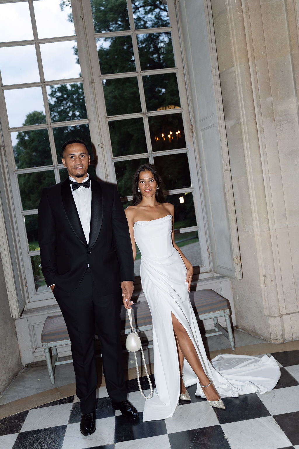 Bride and groom holding hands on a black and white checkered marble floor in a French chateau with tall French windows