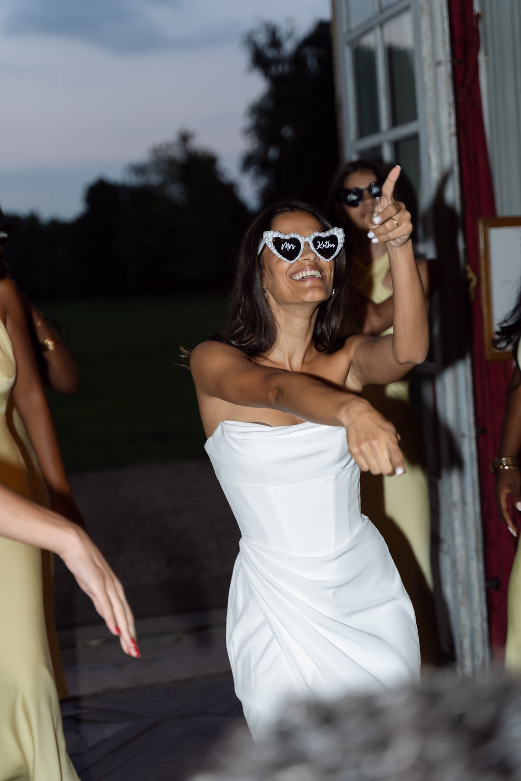 Bride dancing at dusk in short white dress with heart-shaped sunglasses surrounded by bridesmaids in yellow dresses