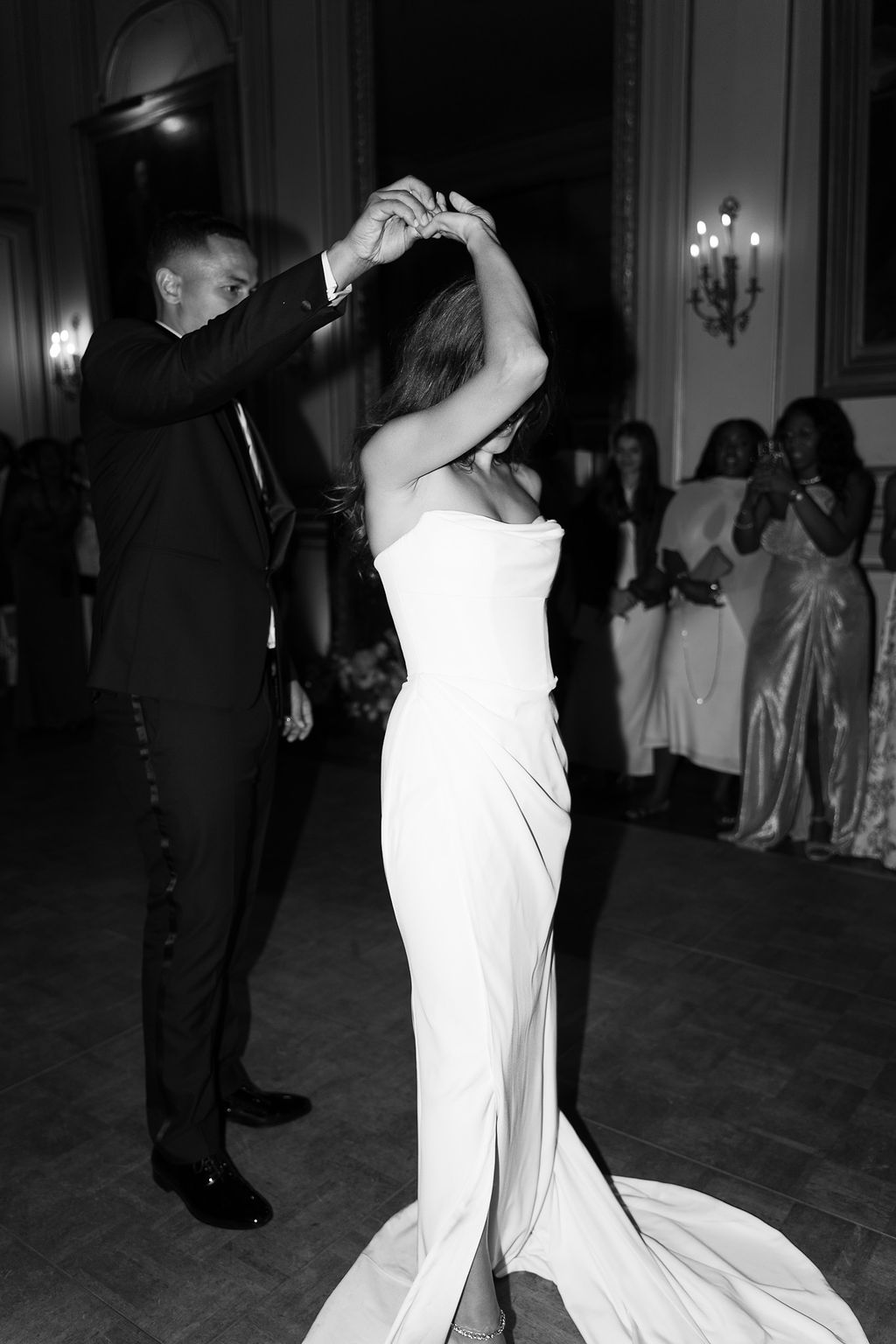 Black and white photo of couple's first dance in grand ballroom with guests watching from the perimeter