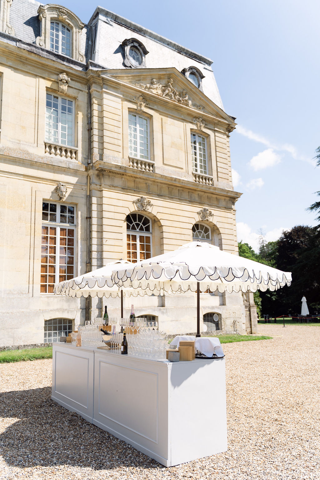 White champagne bar with flutes beneath scalloped umbrella on gravel forecourt of ornate limestone chateau