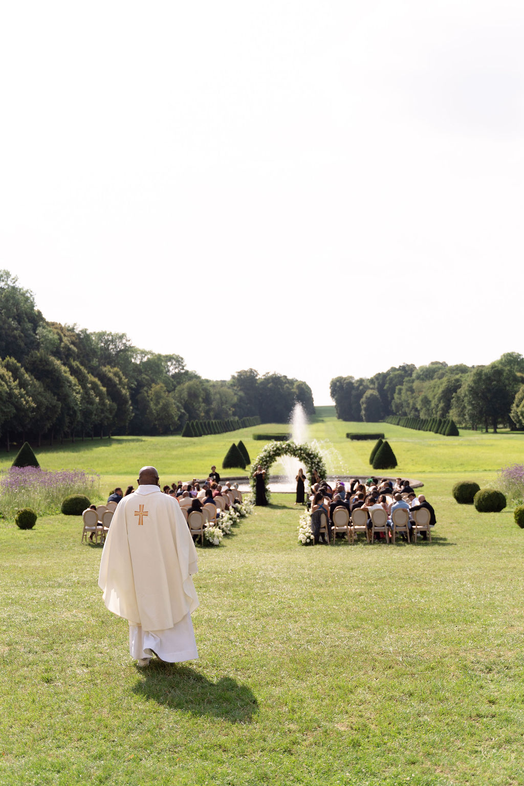 Sixty guests at formal garden ceremony with circular floral arch fountain topiary and priest approaching aisle