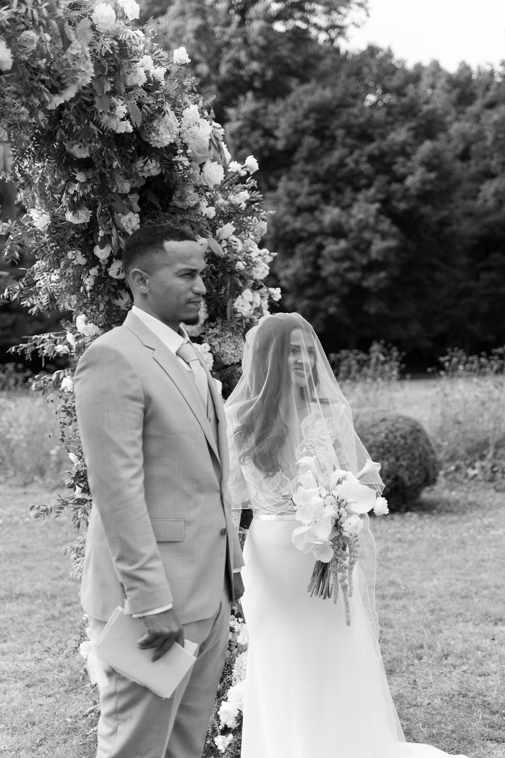Black-and-white portrait of bride and groom standing before a floral arch during outdoor garden ceremony