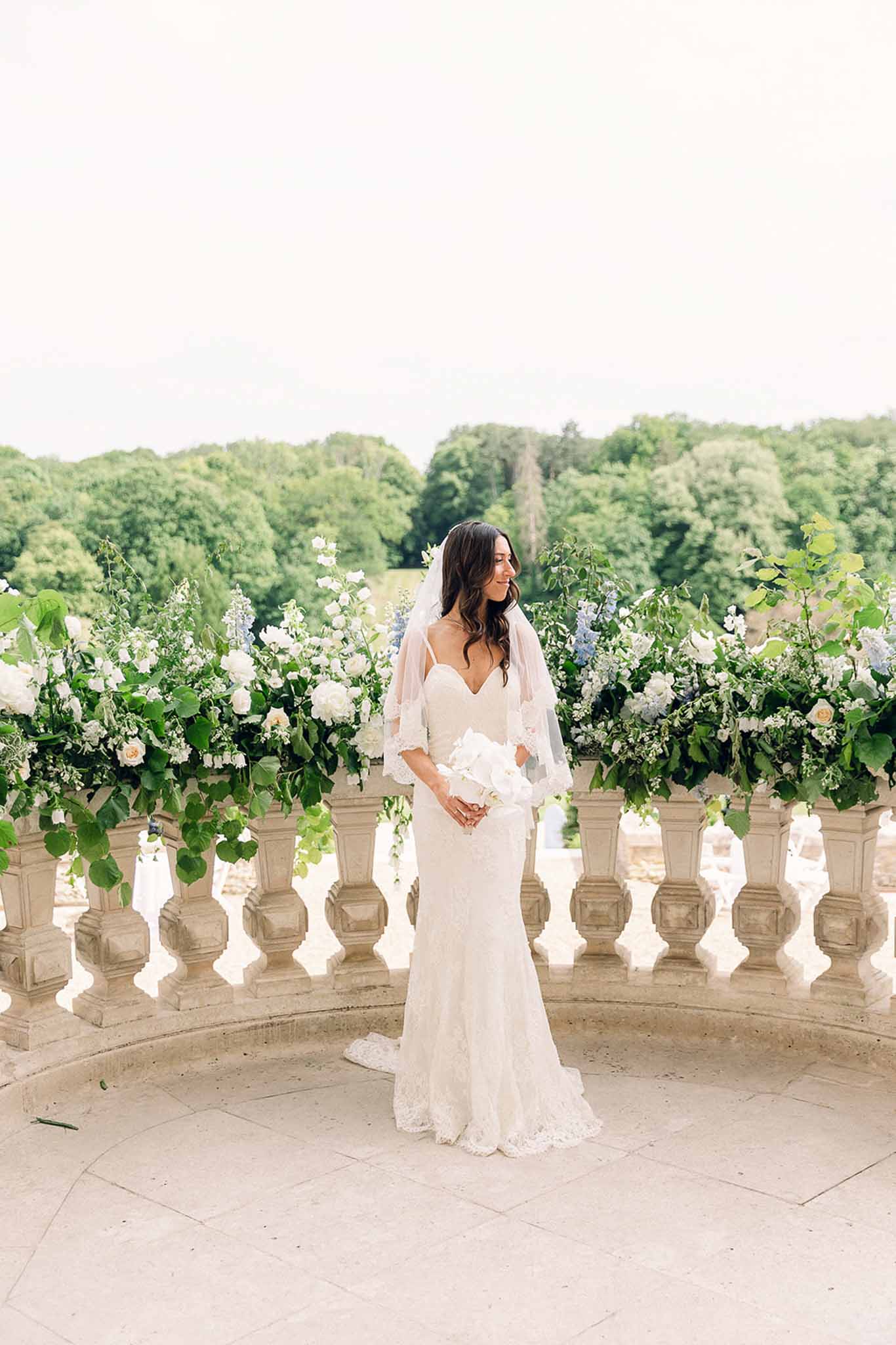 Bride in ivory lace gown with short veil and white bouquet beside stone urns of roses and delphiniums