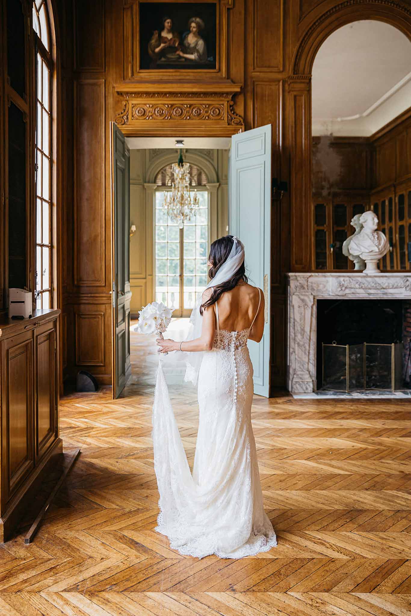 Rear bridal portrait in backless lace gown with orchid bouquet in wood-paneled chateau room