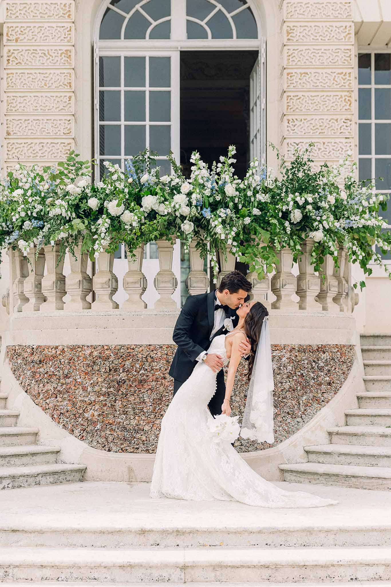 Groom dips and kisses bride on ornate chateau stone staircase lined with white and blue floral urns