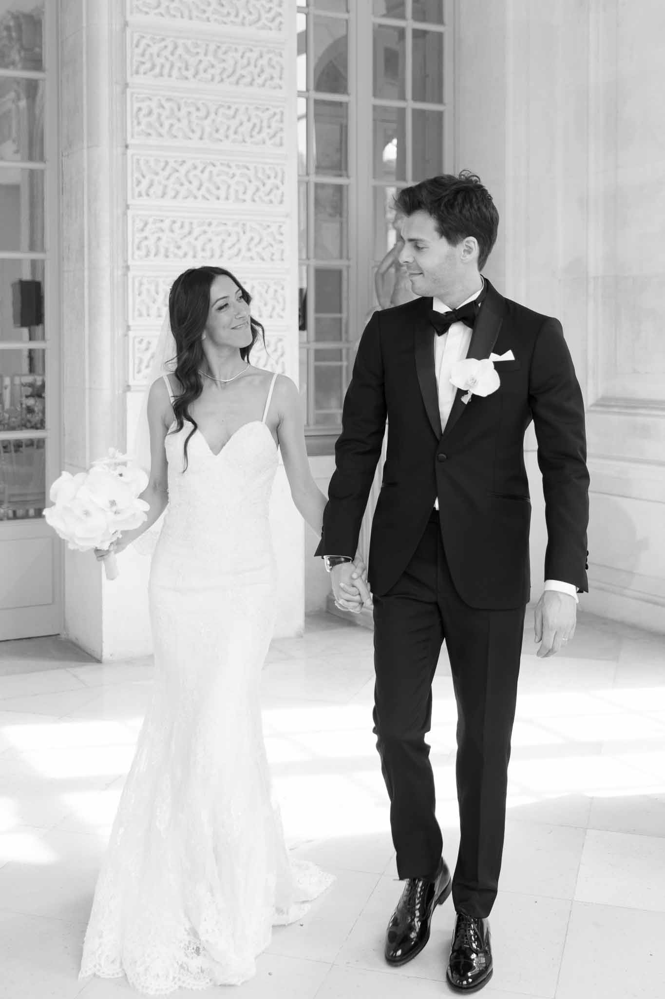 Bride in lace gown with orchid bouquet and groom in tuxedo walk hand in hand on stone terrace in B&W