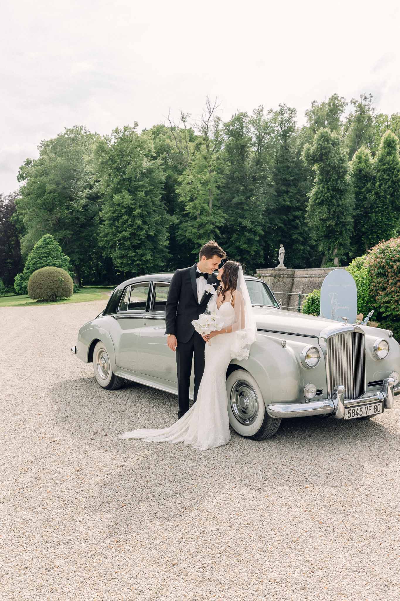 Bride and groom posing before vintage silver Bentley on chateau driveway with topiary backdrop