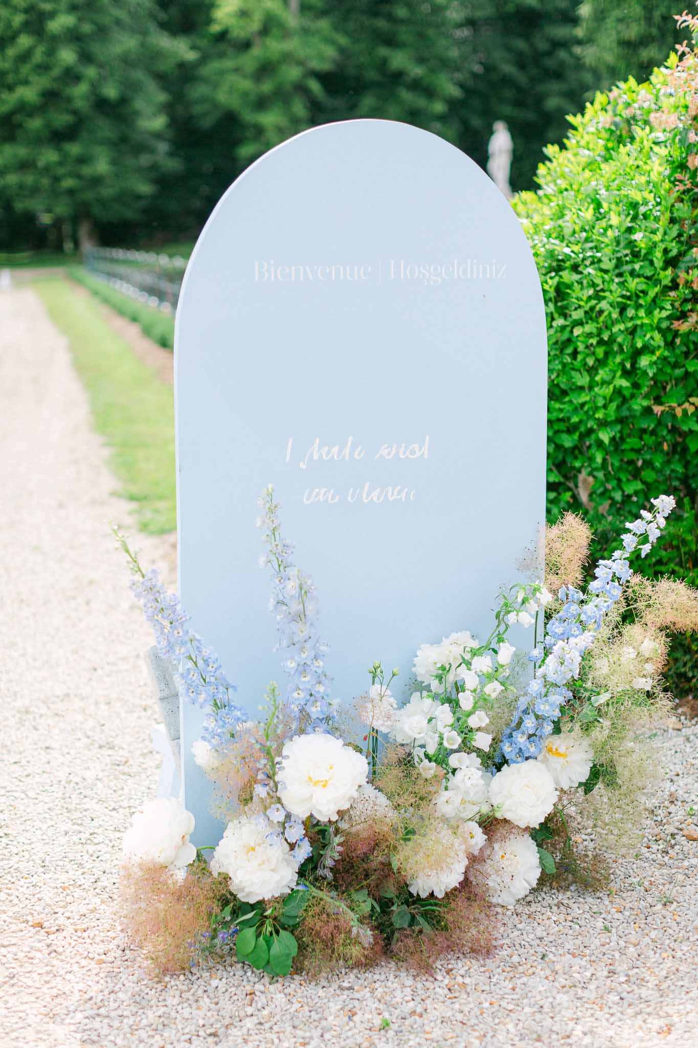 Dusty blue arch welcome sign with ivory peonies blue delphiniums and smoke bush foliage on gravel