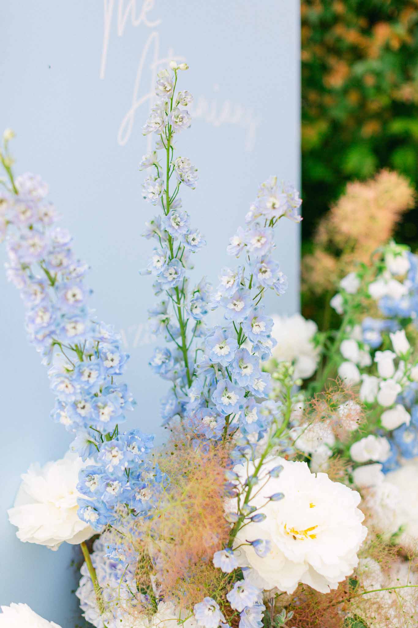 Blue delphinium and white rose arrangement at base of powder blue calligraphy welcome sign outdoors